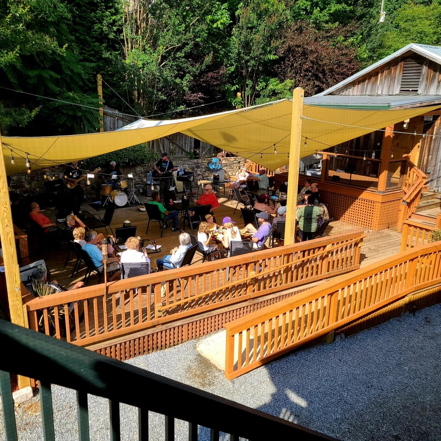 Outdoor deck with wooden railings, yellow sun shades, and string lights, where a live music band performs for an audience seated in chairs and at tables, surrounded by trees and a wooden building.