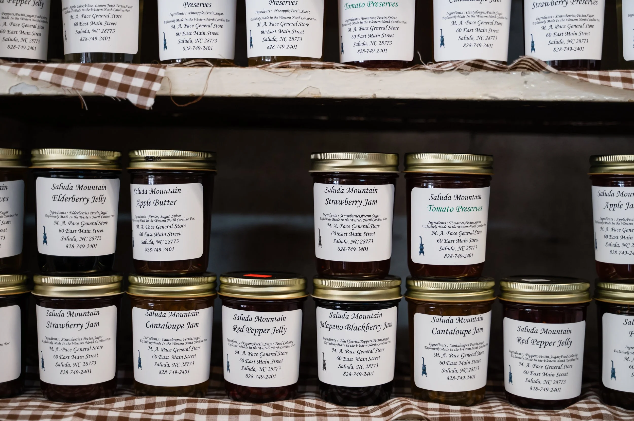 Shelf with jars of homemade jams and preserves, including strawberry jam, apple butter, cherry jelly, and tomato preserves. Each jar has a label with the brand name 'Saluda Mountain' and the store's address in Saluda, North Carolina.