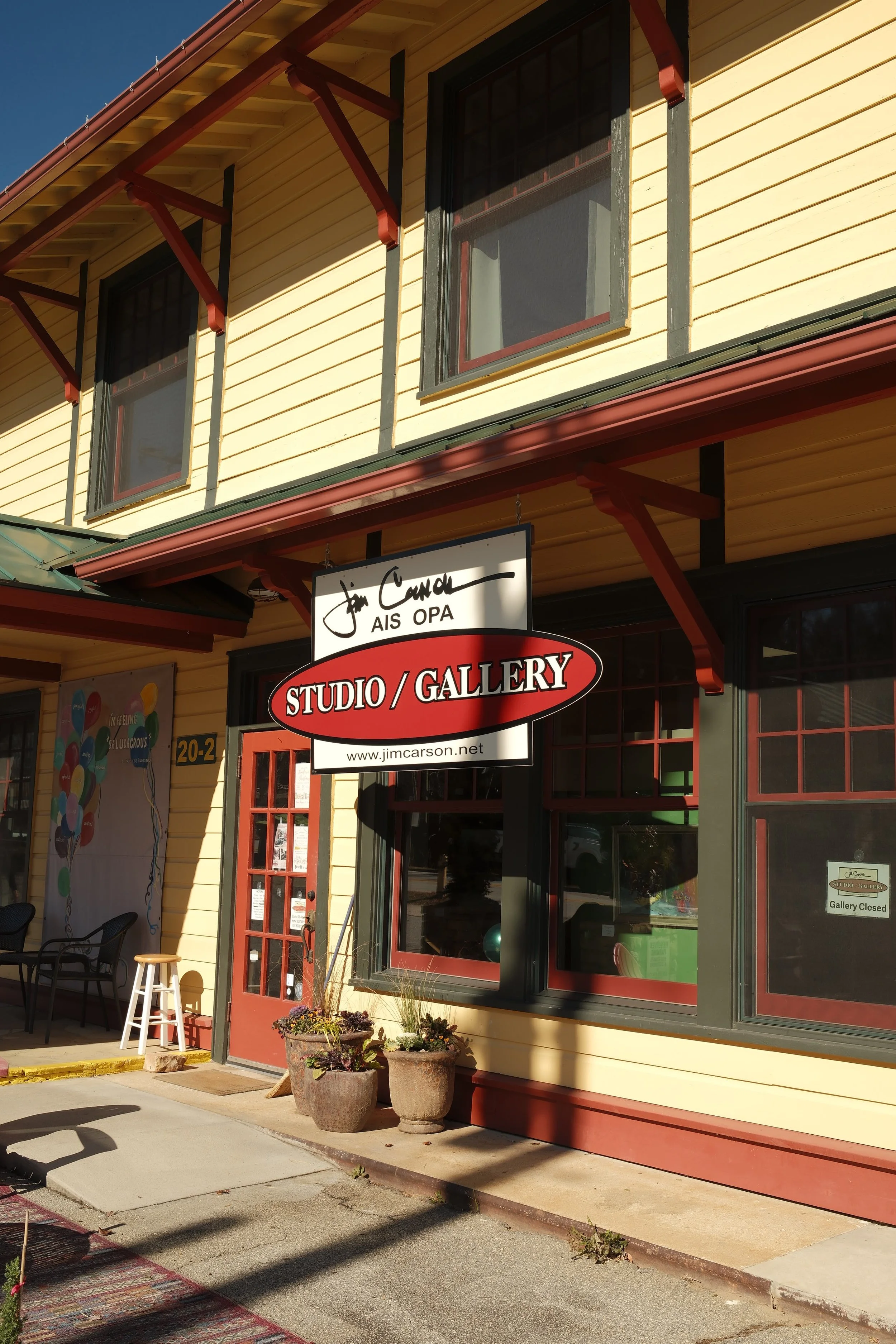 Exterior view of a yellow building with red trim, a sign for Jim Carson Studio/Gallery, outdoor seating with chairs, potted plants near the entrance, and a closed gallery door with a sign.