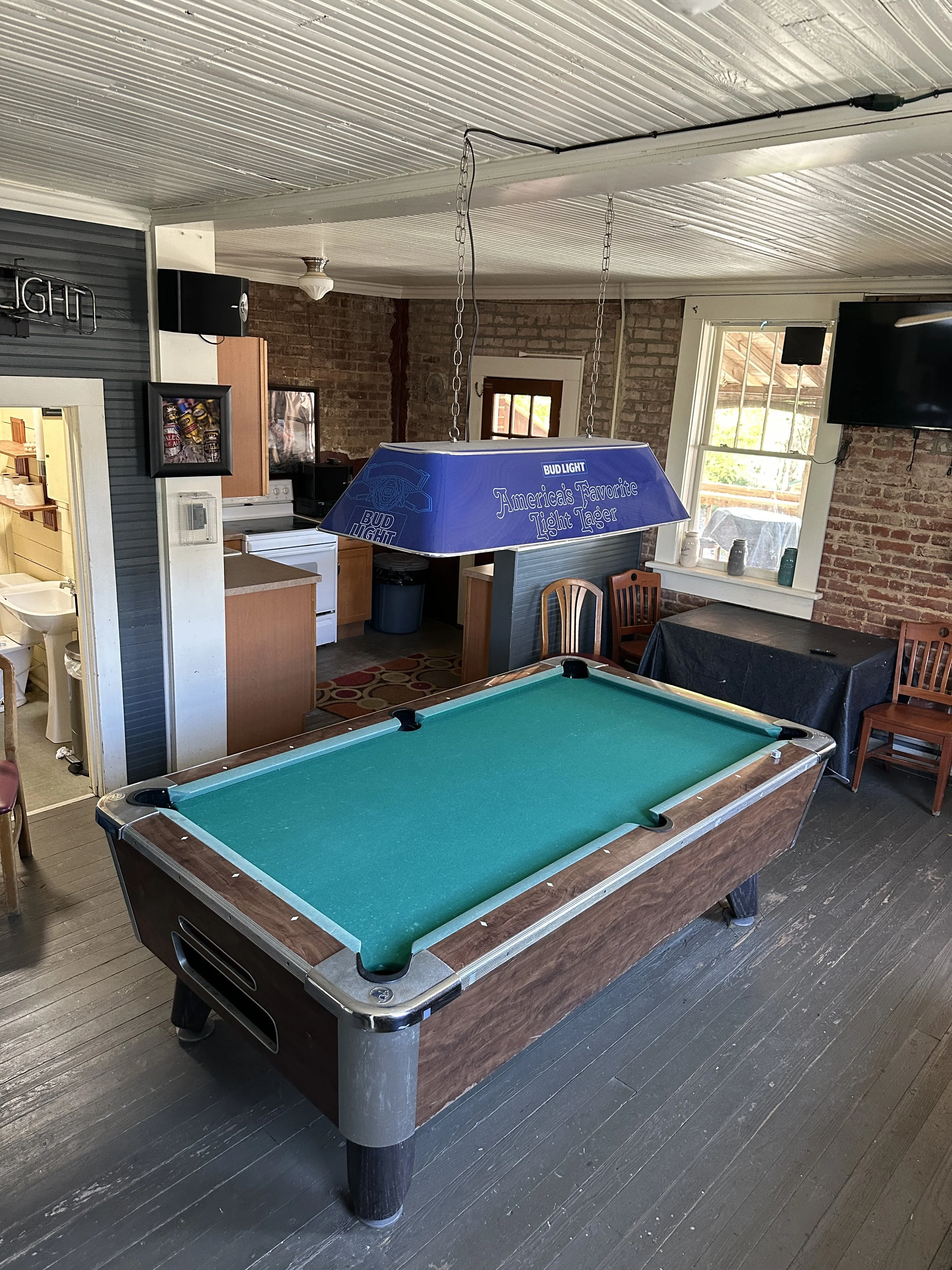 Pool table with green felt in a room with brick walls, a window, and a hanging Bud Light sign-shaped light fixture.