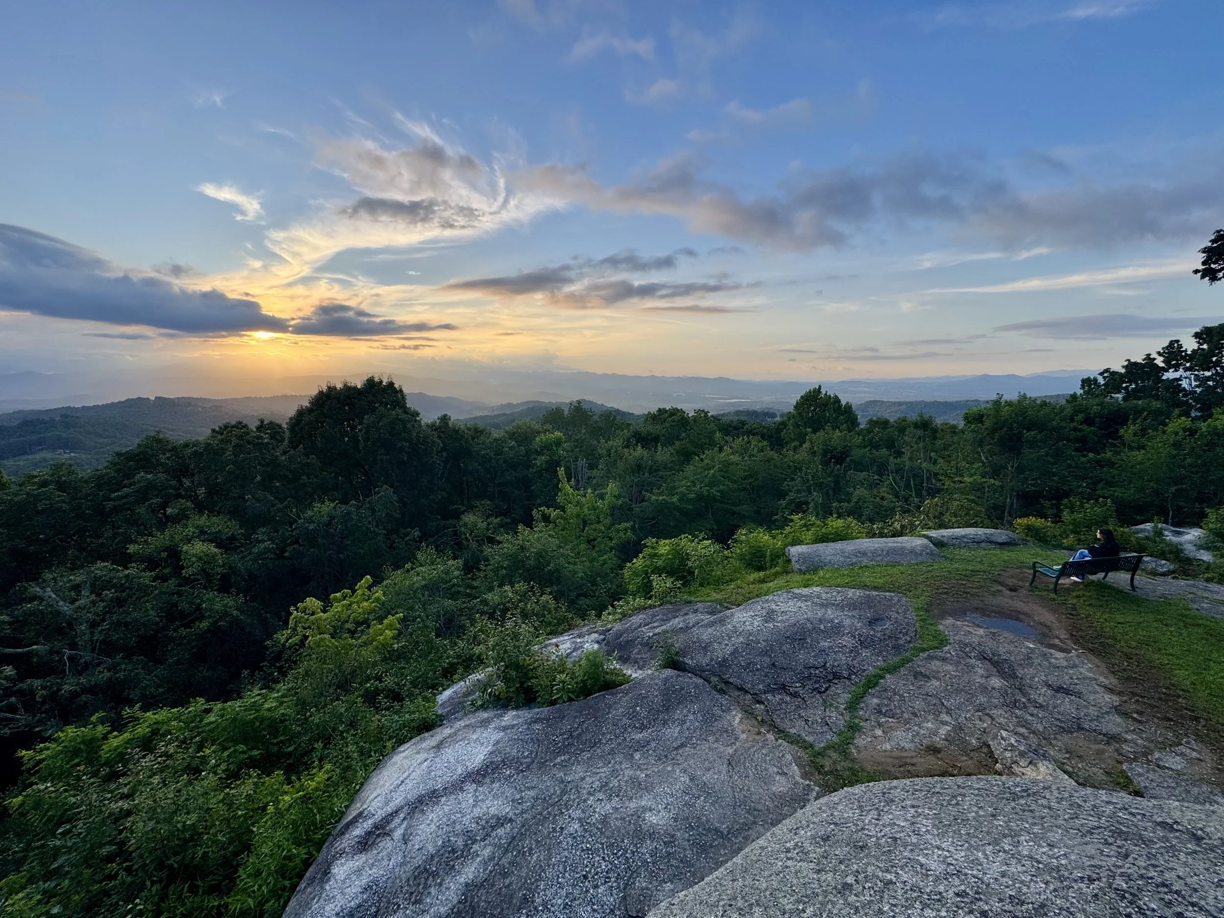 A scenic view of a sunset over rolling hills with a sky partly cloudy, green trees in the foreground, and a person sitting on a bench near rocks.