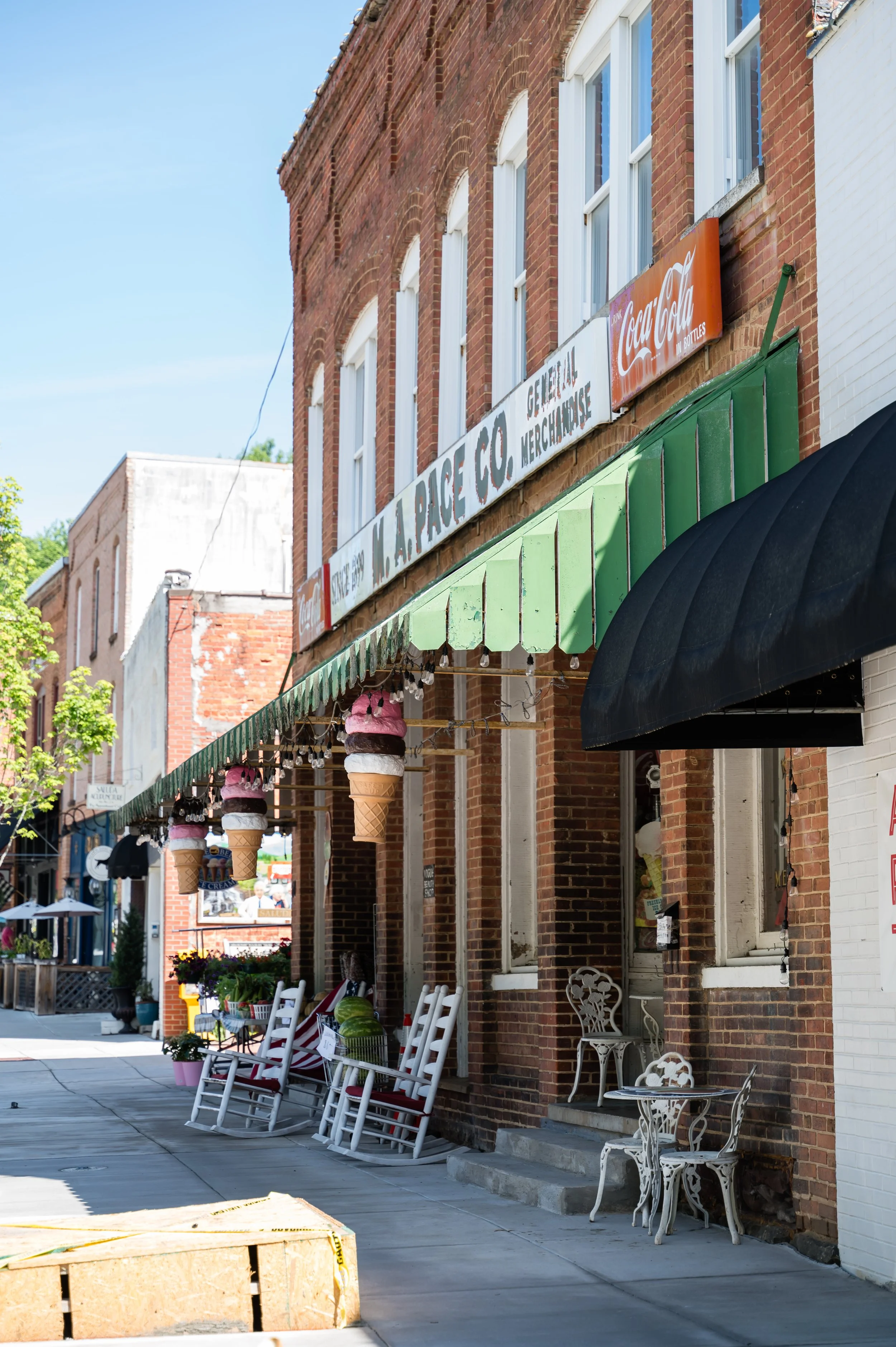 Downtown storefronts with outdoor seating, ice cream cone decorations, and vintage chairs under green awnings on a sunny day.