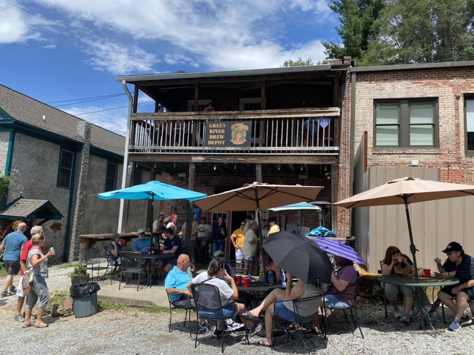 People gathered outdoors near a brick building with a sign that reads 'Green River Brew Depot.' Some are sitting under umbrellas, while others are standing and socializing.
