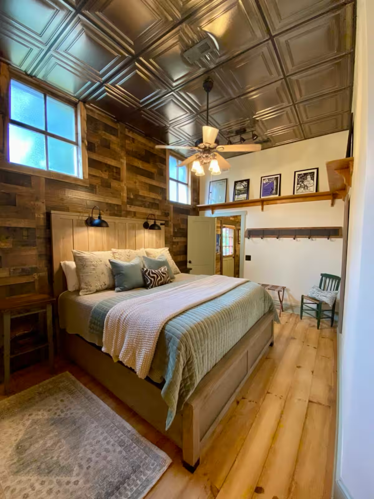 A bedroom with wooden walls and a wooden floor, featuring a large bed with multiple pillows, a ceiling fan with lights, and framed pictures above a shelf on the wall.