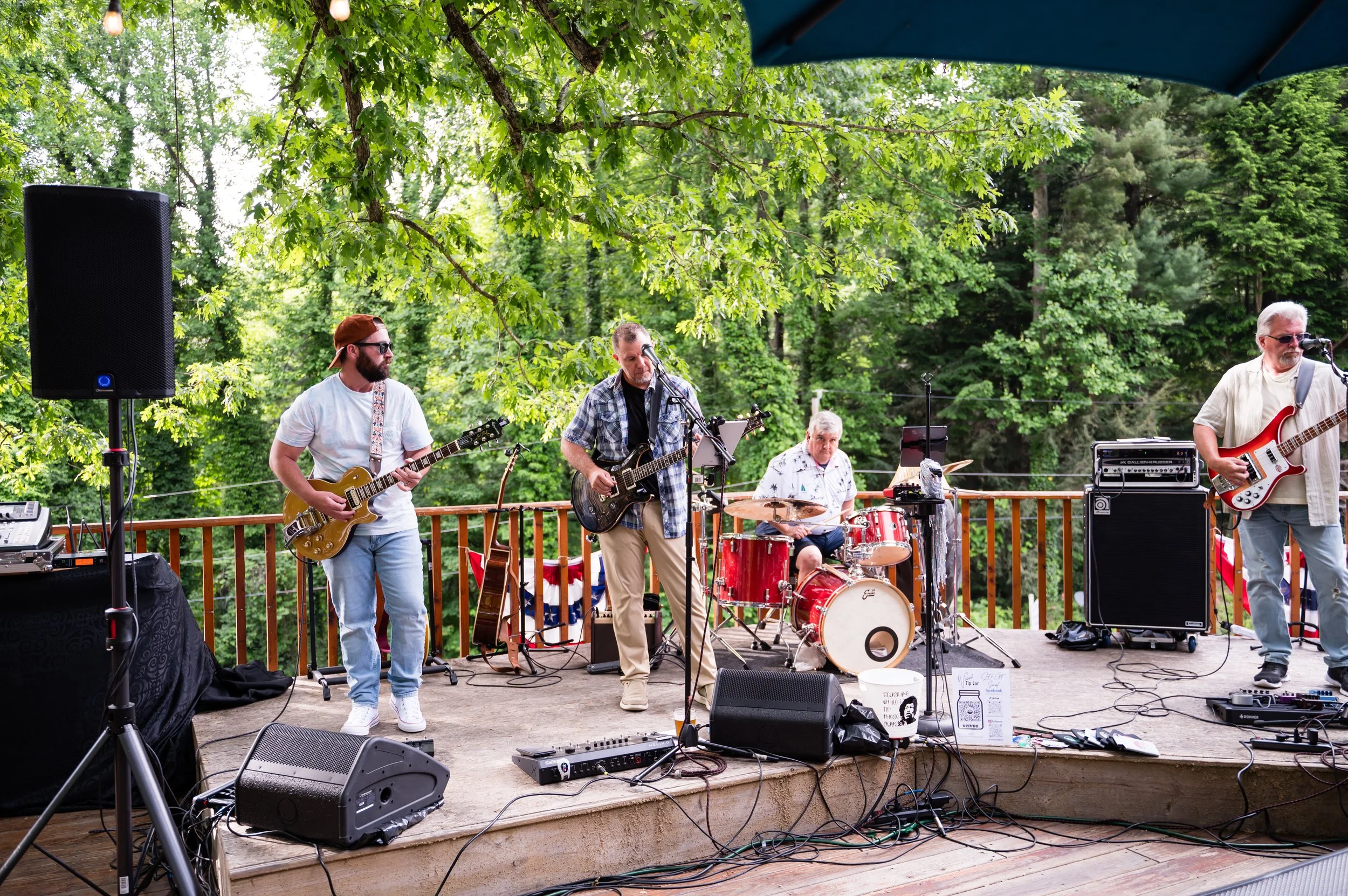 Four musicians performing outdoors on a wooden stage surrounded by green trees, with musical equipment including guitars, drums, and speakers.