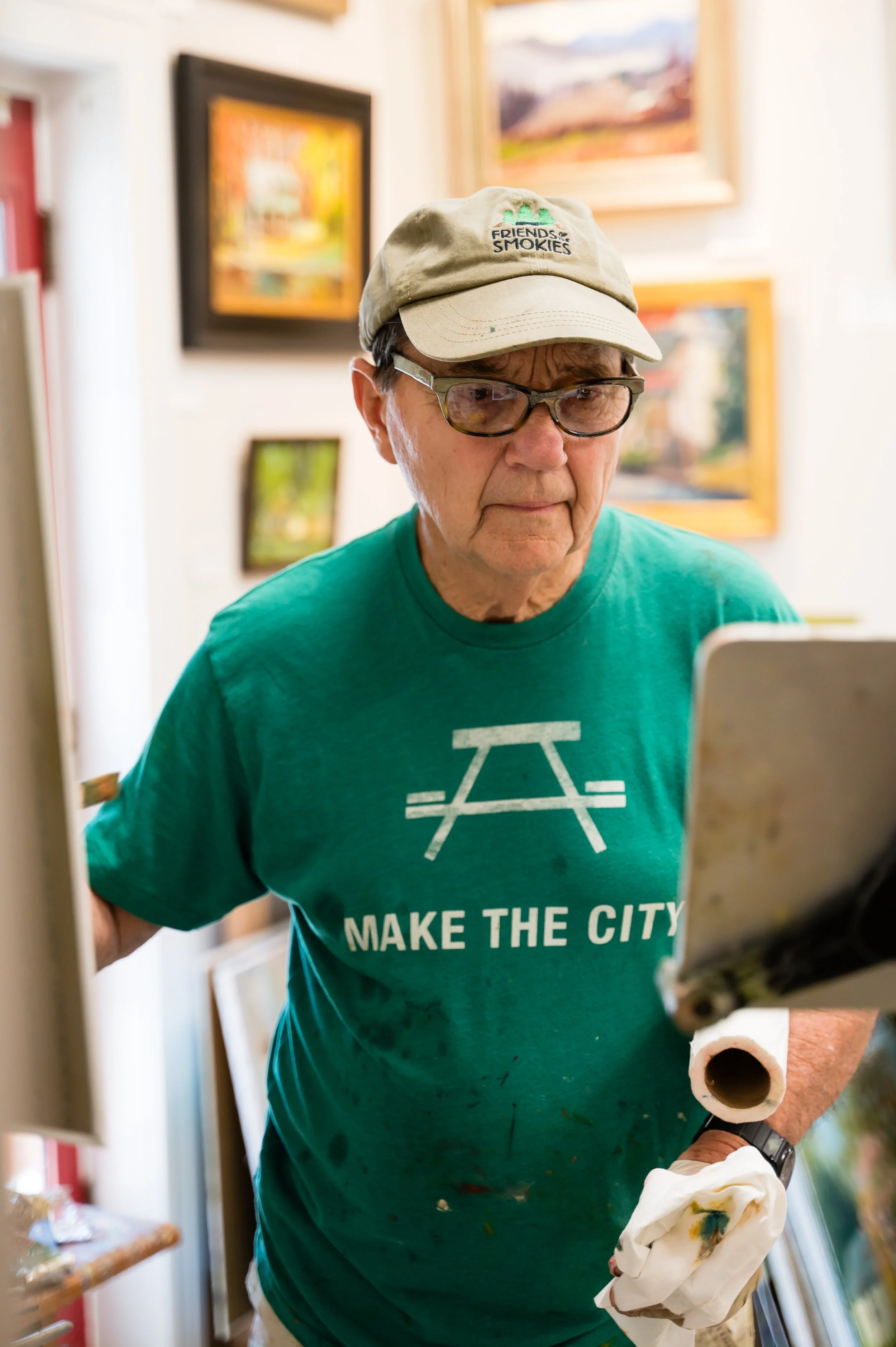 An older man with glasses and a beige hat with the logo 'Friends of Smokies' on it, wearing a green T-shirt that says 'Make the City' with a graphic, is holding a paint roller and a cloth while painting or working on an art project in a room with col