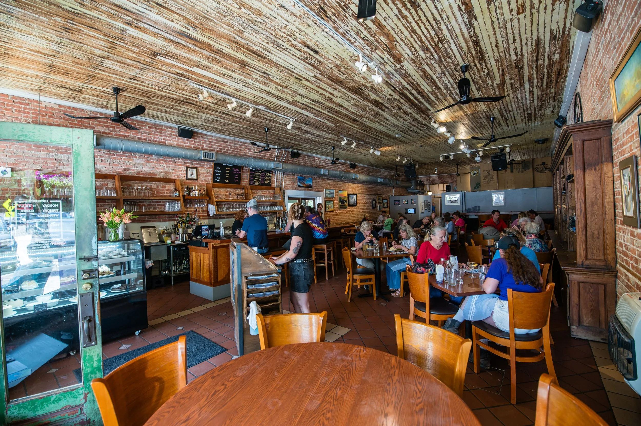 Interior of a busy restaurant with wooden tables and chairs, exposed brick walls, and a wooden ceiling. Customers are dining, and staff are working behind the counter. Artwork decorates the walls.