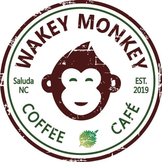 Logo for Wakey Monkey Coffee Cafe with a smiling cartoon monkey face, green and brown text, established 2019 in Saluda, North Carolina, and a green leaf graphic.