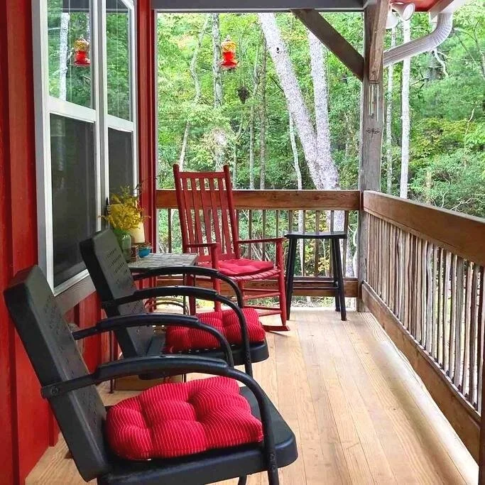 View of a wooden porch with three chairs with red cushions, small black table, and yellow flowers, overlooking a lush green forest.