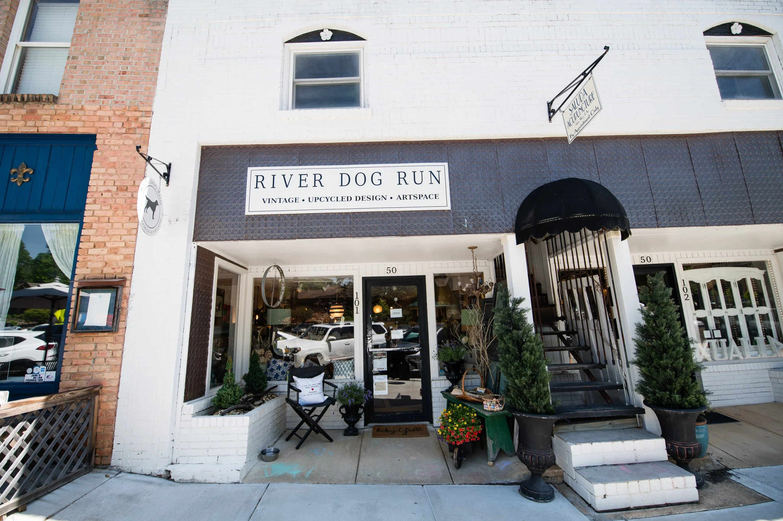 Storefront of River Dog Run, a vintage, upcycled design art space, with a white brick exterior, large window, and entrance decorated with plants and outdoor furniture.