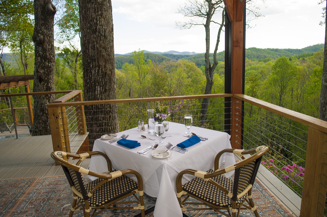 Elegant outdoor dining table set with blue napkins, glassware, and flowers, on a deck overlooking a lush green forested landscape.