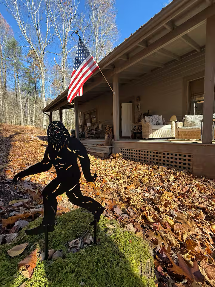 A metal silhouette statue of a Bigfoot or Sasquatch figure on a moss-covered hill in front of a house with a porch. An American flag is hanging from the porch roof. The yard is covered with fallen autumn leaves, and there are bare trees and a clear blue sky in the background.