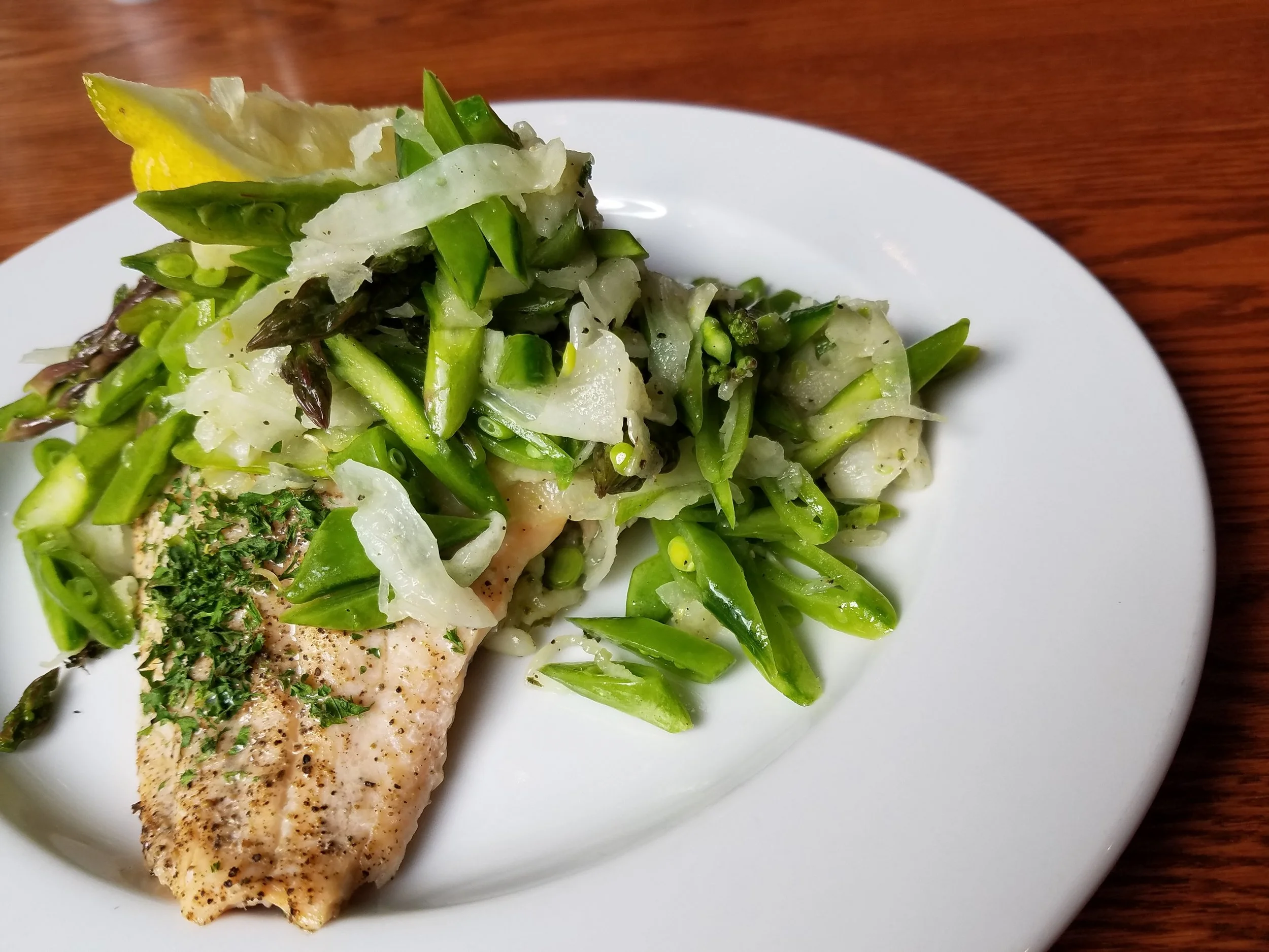 A white plate with a portion of cooked fish topped with chopped herbs, served with mixed cooked green vegetables including asparagus, green beans, and snap peas.