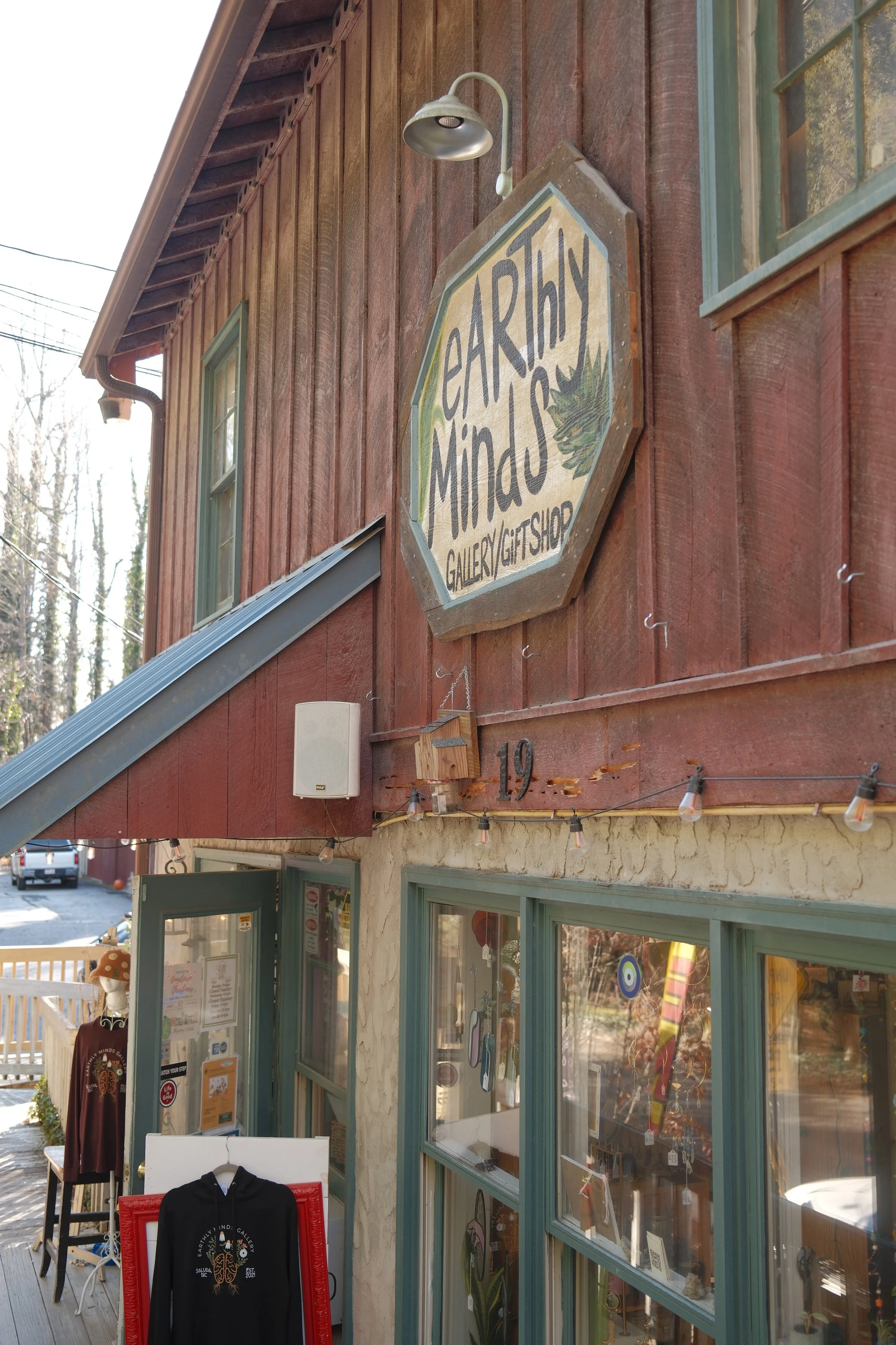 Exterior of a rustic gift shop called 'Earth + Mindy Gallery Gift Shop' with wooden siding, large windows, a mountain landscape sign, and outdoor merchandise display including a hoodie and a manekin.