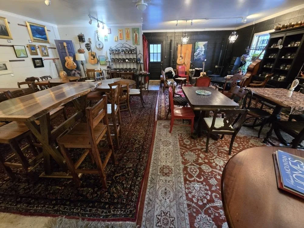 A cozy room decorated with guitars on the wall, framed artwork, and various furniture including tables, chairs, and a bookshelf filled with items. The room has patterned area rugs and a window with curtains.