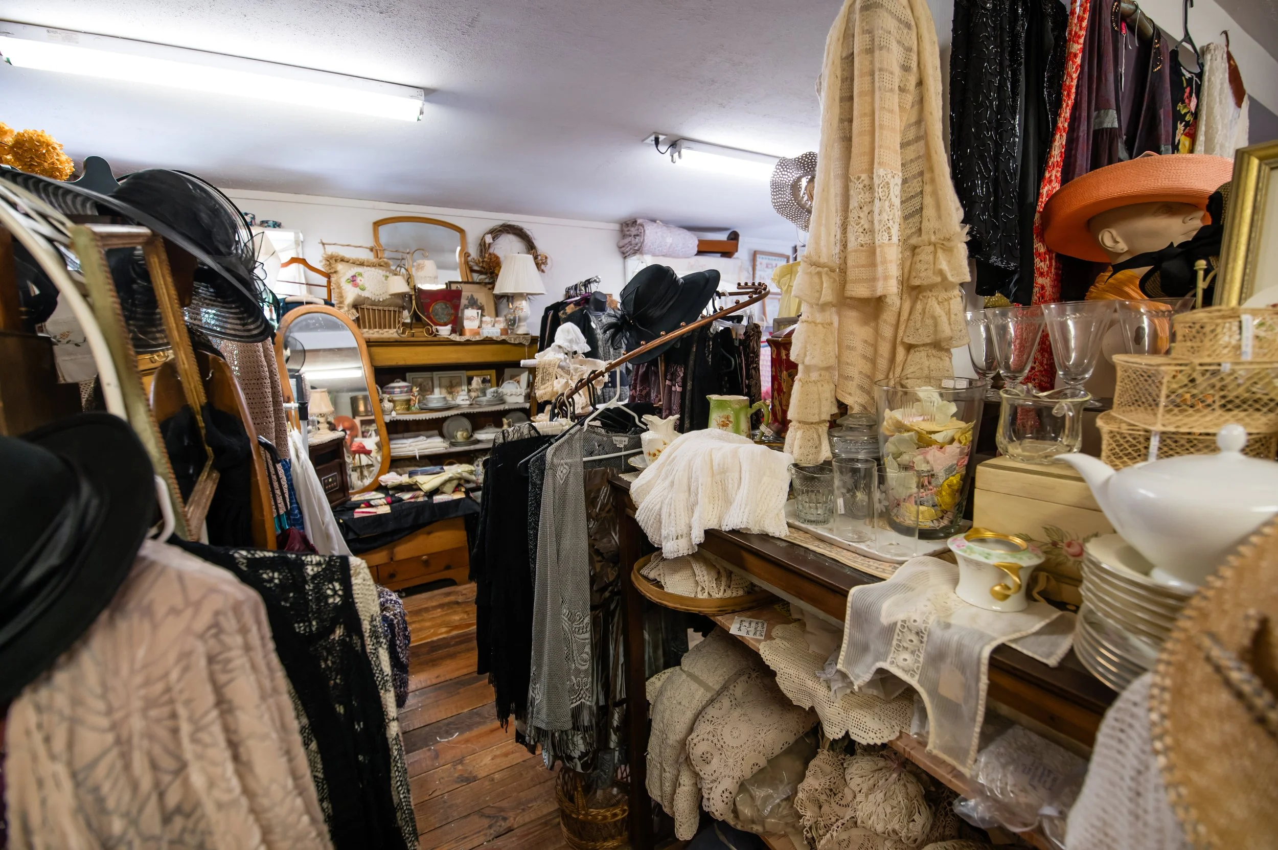 A cluttered thrift store or antique shop filled with clothes, hats, vintage decor, glassware, and framed pictures. The space has wooden floors and fluorescent lighting.