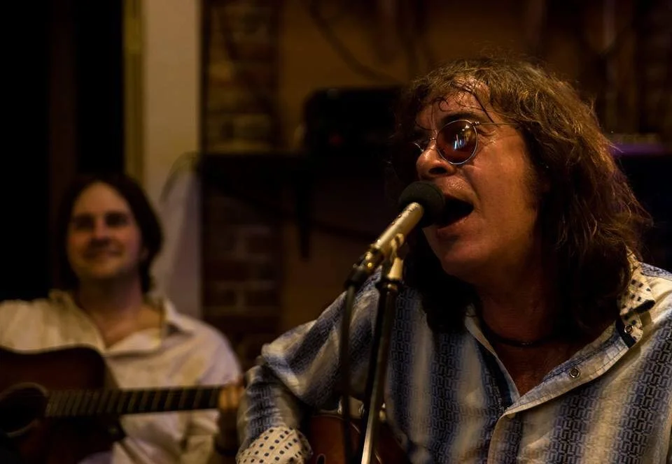 Two men playing guitars and singing into microphones in a dimly lit room with brick walls.