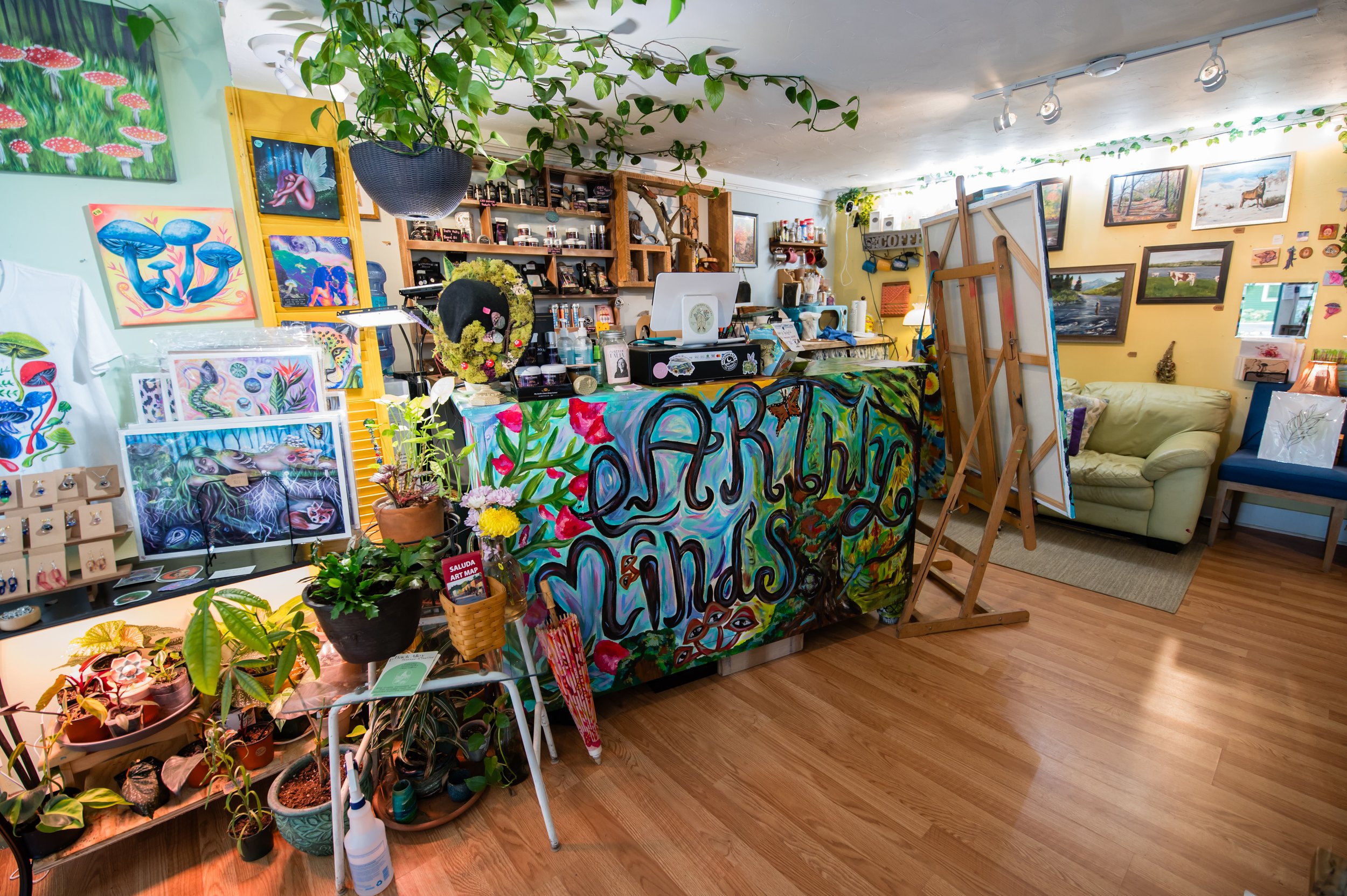 Interior of an art shop decorated with vibrant paintings of mushrooms, animals, and fantastical scenes. There are shelves with art supplies, potted plants, and a counter covered with colorful artwork and merchandise. The wall on the right features fr
