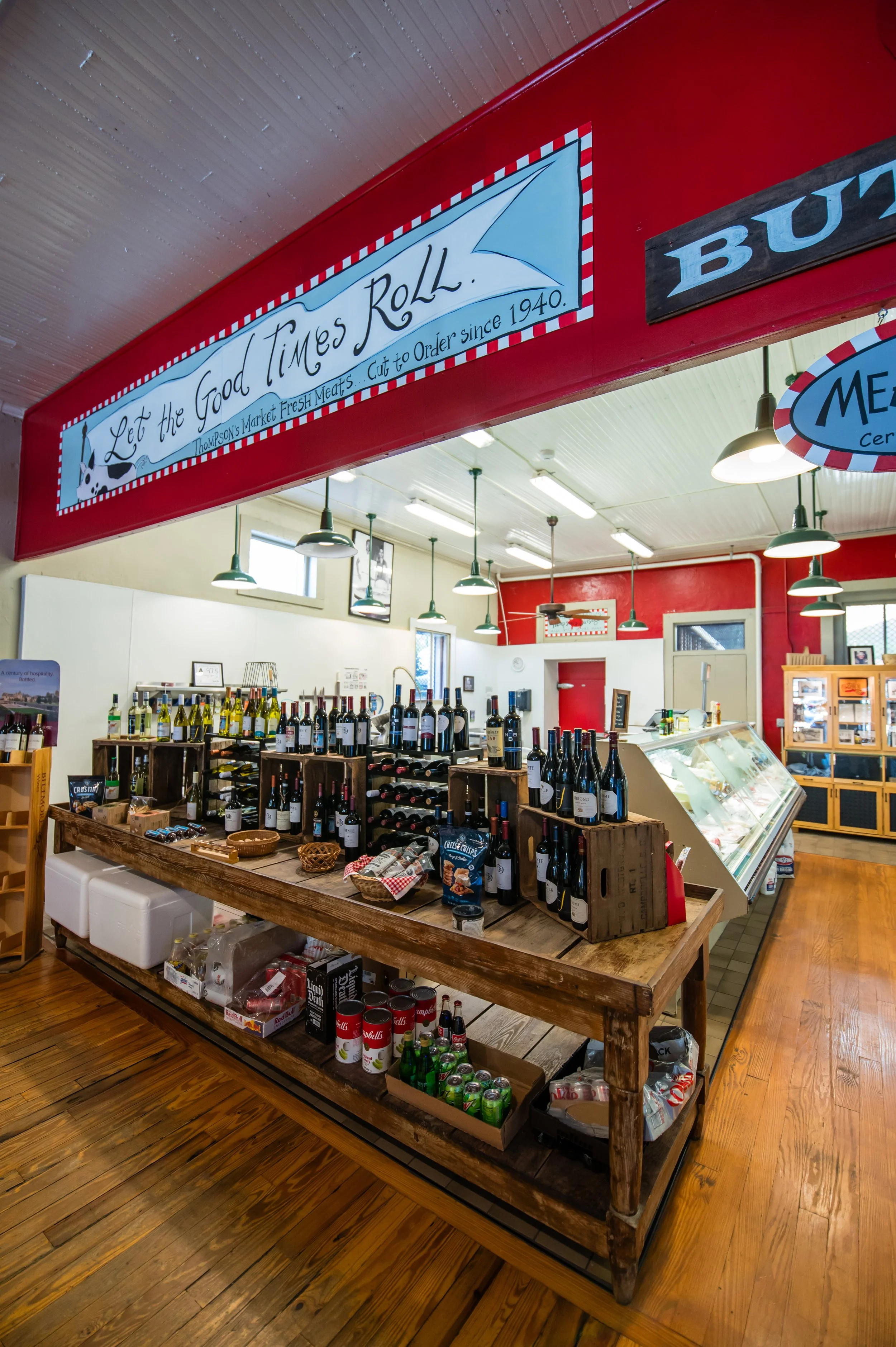 Interior of a store or market with a wooden table displaying various bottles of wine, canned goods, and snacks. Hanging lights and a red wall with signs, including one that says "Let the Good Times Roll" and another partially visible sign that says "