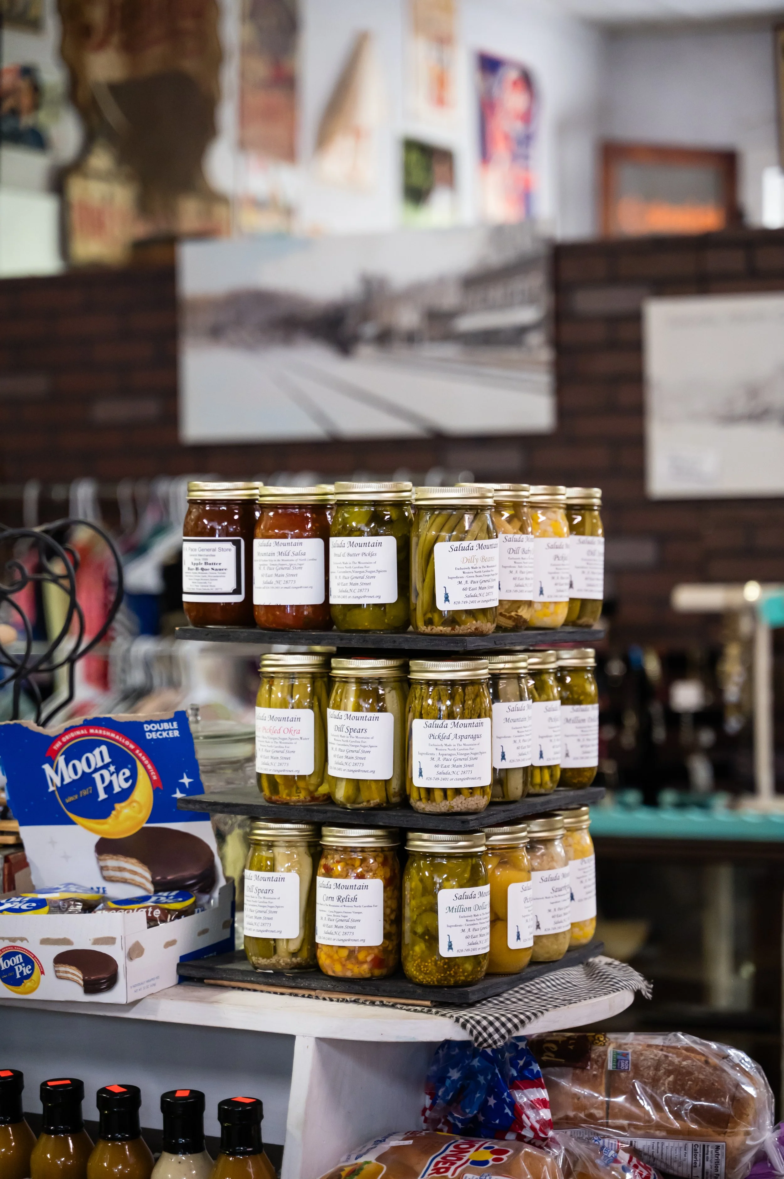 Display shelf with jars of pickled vegetables and preserves in a store, with artwork and posters on the wall in the background.
