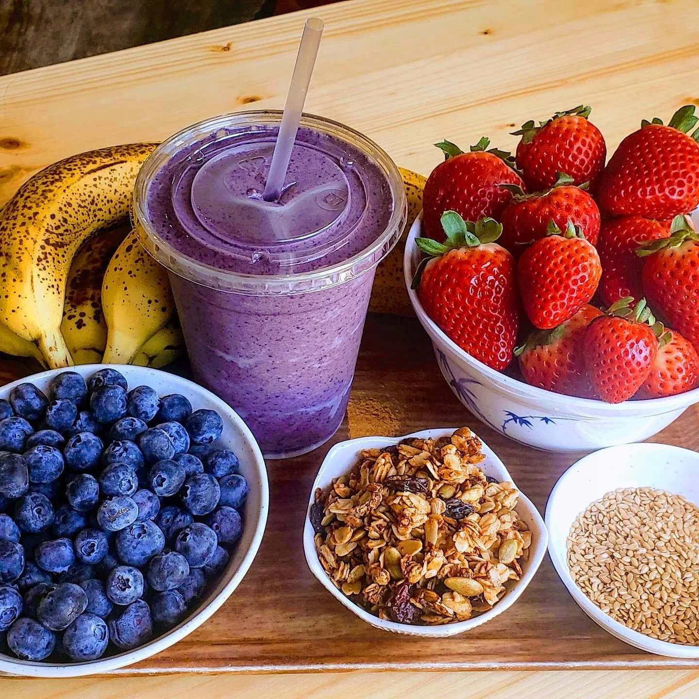 A wooden table with bowls of blueberries, granola, sesame seeds, strawberries, bananas, and a purple smoothie with a straw.