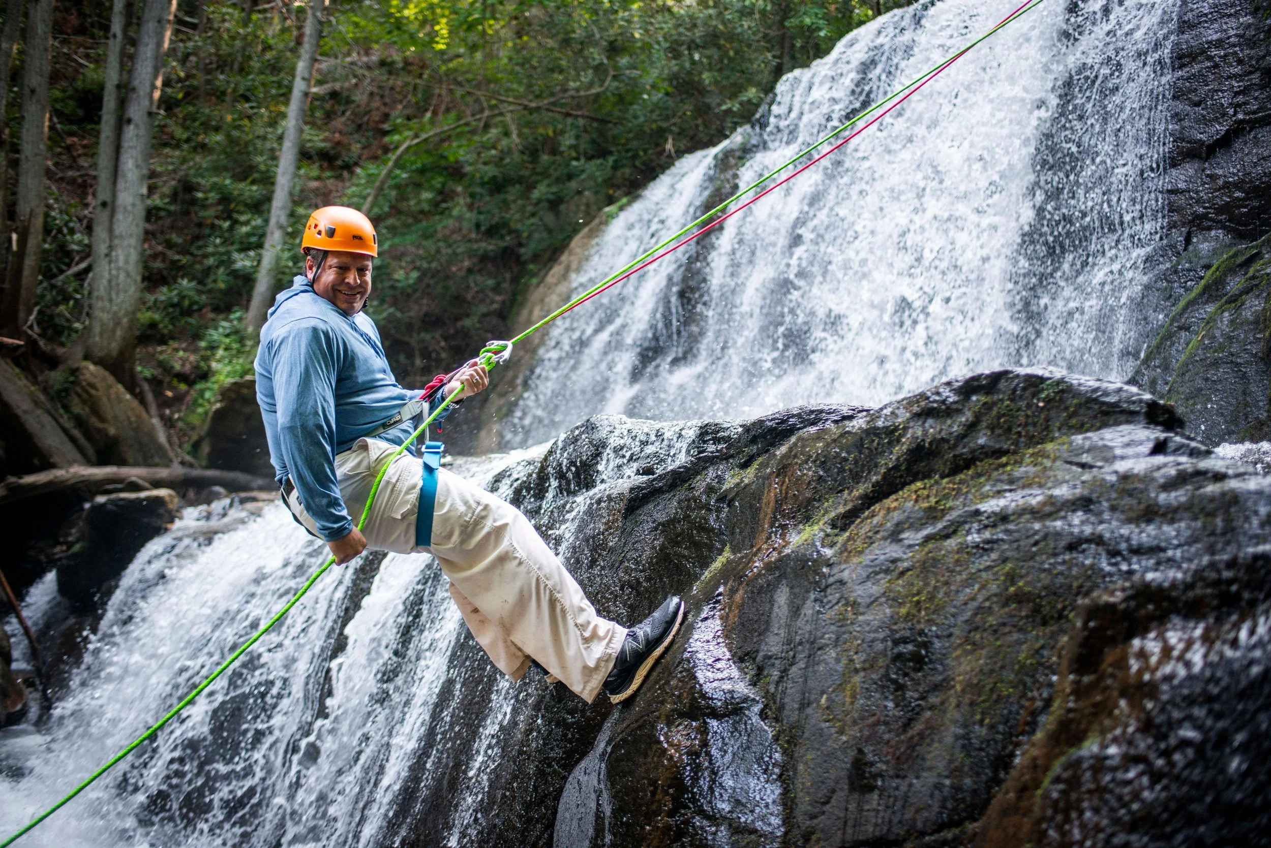 A man rock climbing near a waterfall, wearing an orange helmet and a blue shirt, with a harness and climbing rope.