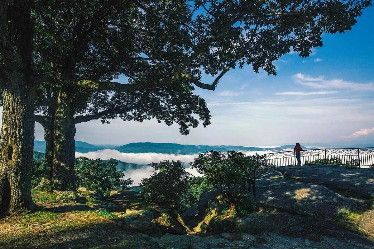 A person standing on a rocky overlook looking at a distant mountain range and low-hanging clouds, with large trees on the left and a blue sky overhead.