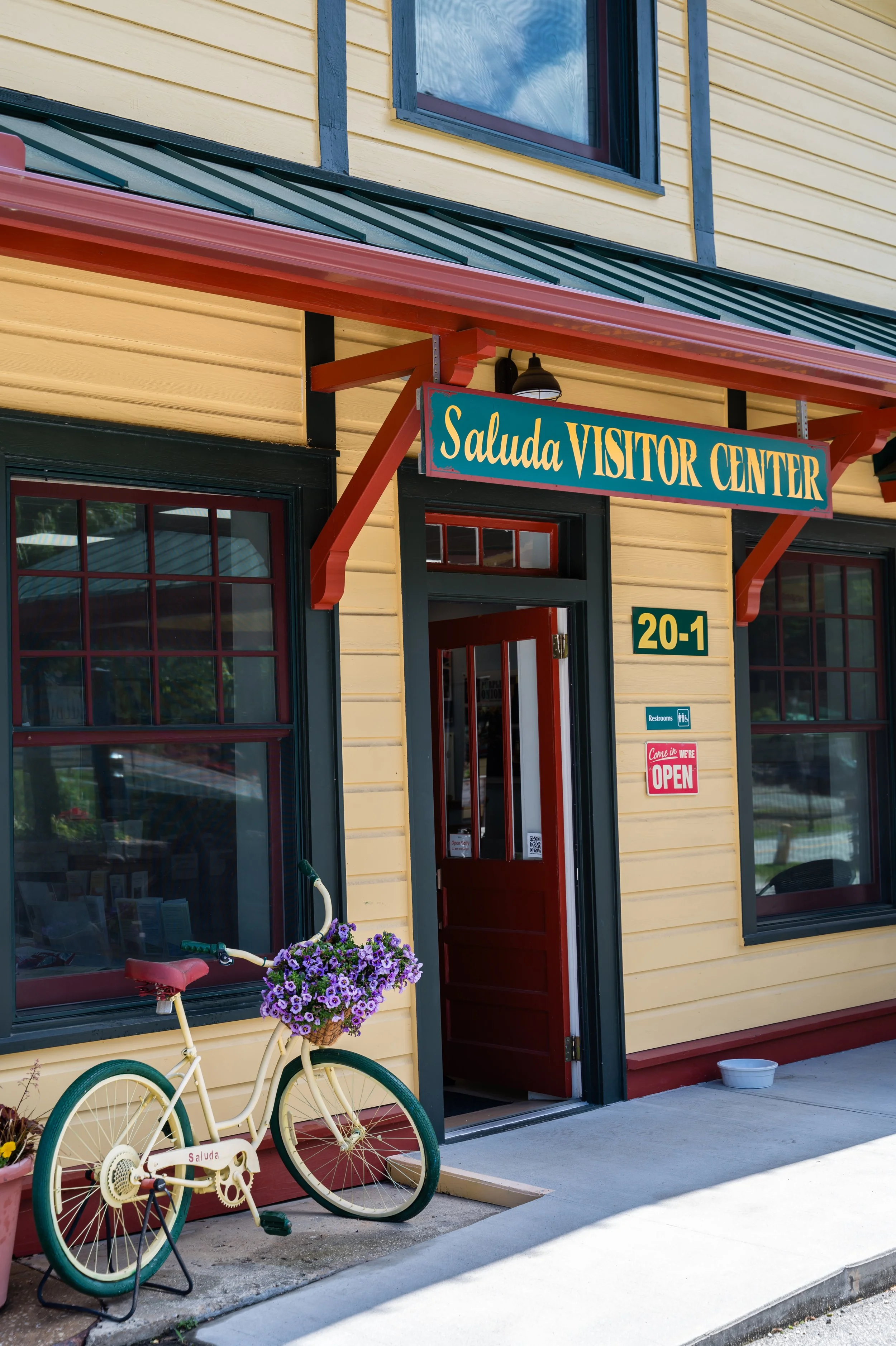 The exterior of the Saluda Visitor Center building with yellow siding, red door and window frames, a yellow bicycle with a basket of purple flowers parked outside, and a sign indicating the visitor center. There are two signs on the wall, one indicating restrooms and another indicating the facility is open.