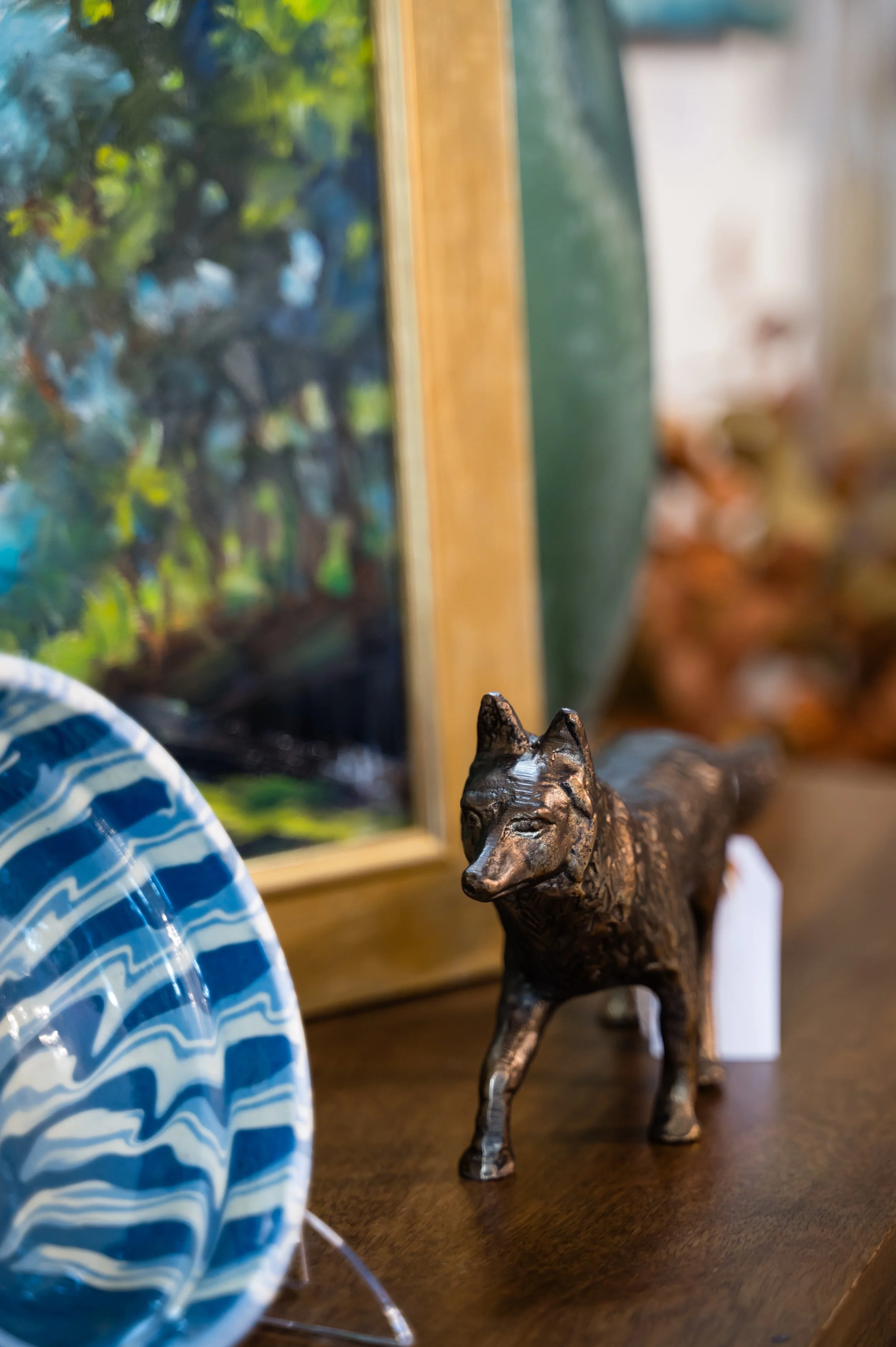 A small bronze-colored wolf figurine on a wooden surface with a blurred painting of trees in the background and a blue and white string ball partially visible on the left.