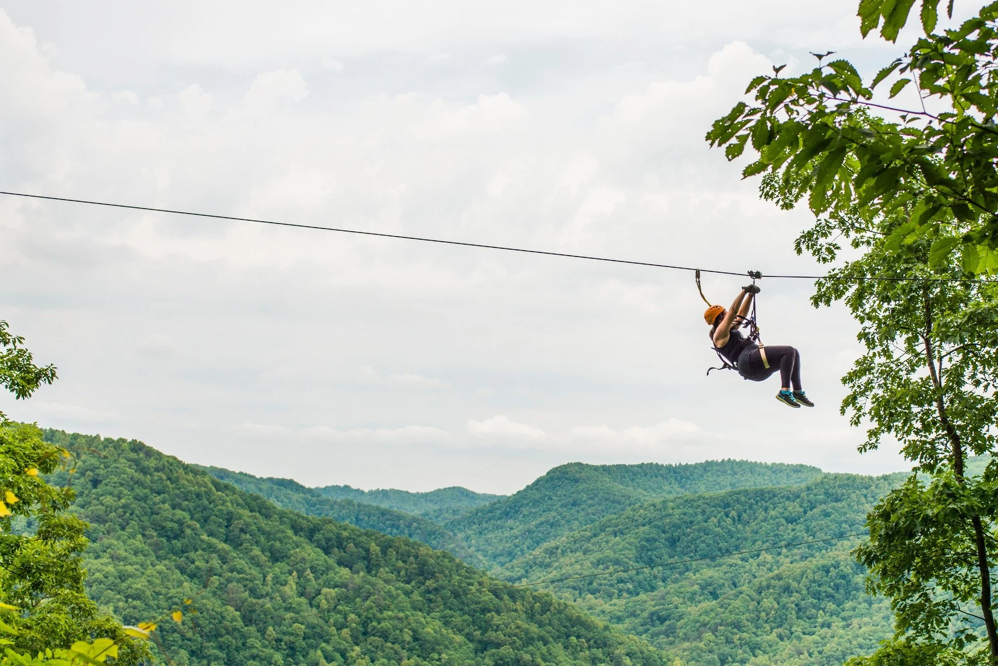 A person wearing a helmet and harness gliding on a zip line through a lush green forested mountain landscape under cloudy sky.