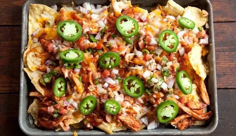 Baked nachos topped with shredded cheese, chopped onions, chopped tomatoes, sliced green jalapeños, and shredded chicken.