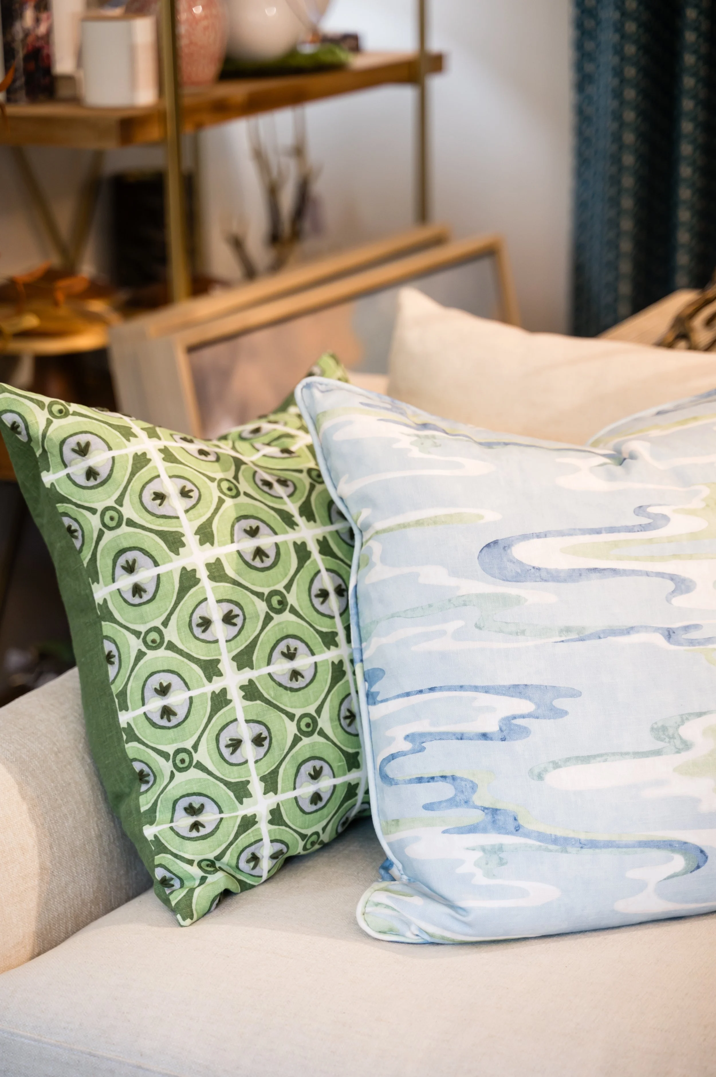 Close-up of two throw pillows on a beige sofa, one with a green and white geometric pattern, the other with a light blue and white abstract pattern. In the background, a wooden shelf with decorative items is visible.