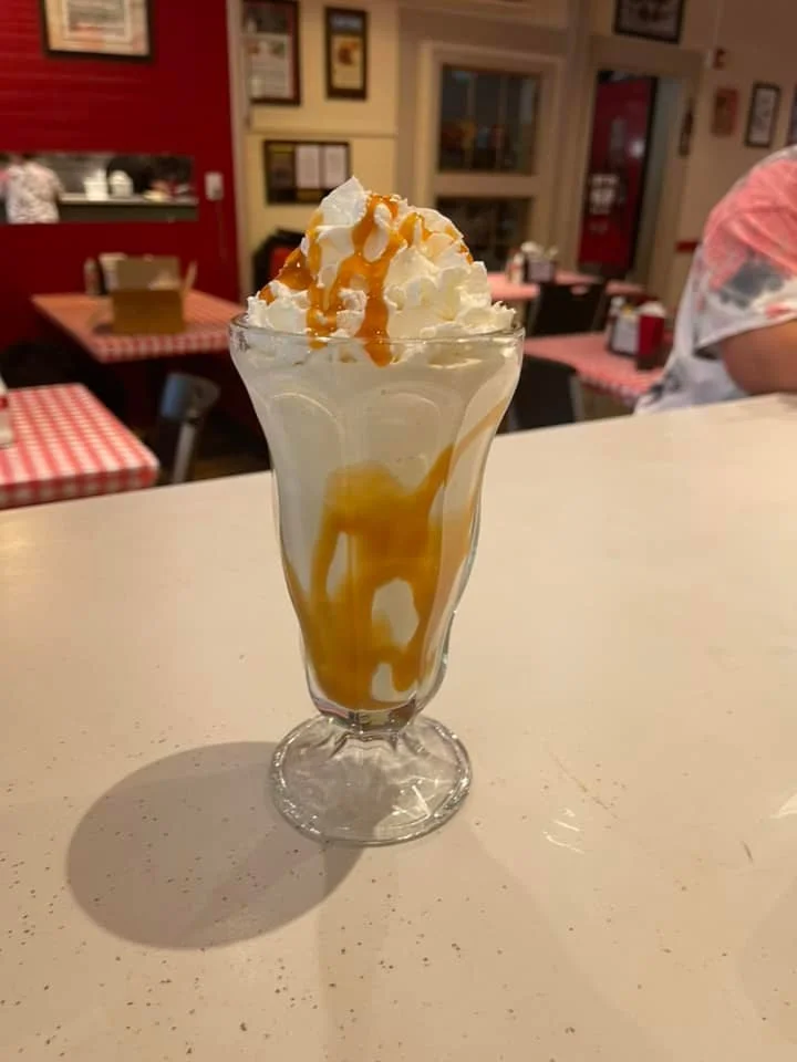 A tall glass of vanilla ice cream sundae topped with whipped cream and caramel sauce, with caramel sauce drizzled inside the glass.