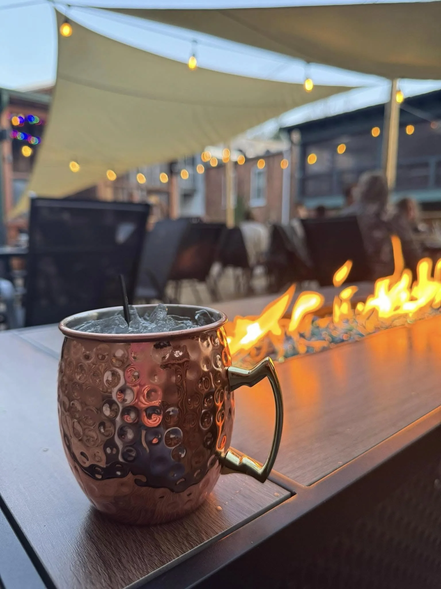 A close-up of a metallic mug filled with ice and a black straw on a table at an outdoor dining area, with string lights and people sitting at tables in the background.