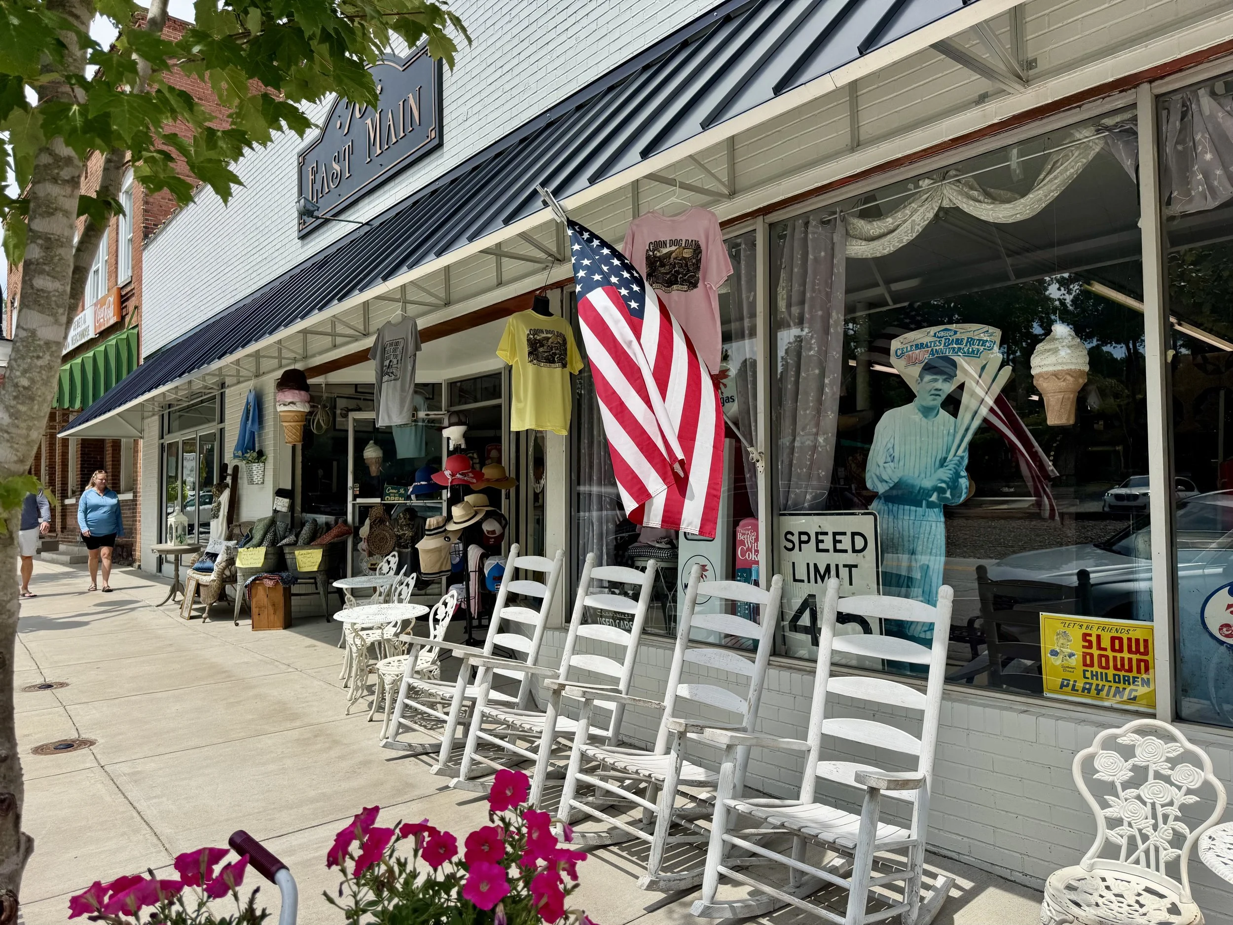 Storefront with white brick wall, black awning, and large display window. Hanging from the awning are colorful T-shirts, an American flag, and a sign with the words 'EAST MAIN'. Outside are white chairs, pink flowers, and a sign indicating a speed limit. Inside the window, a mannequin in a striped shirt and black hat, and a display with ice cream cones and various hats are visible. Pedestrians walk along the sidewalk.