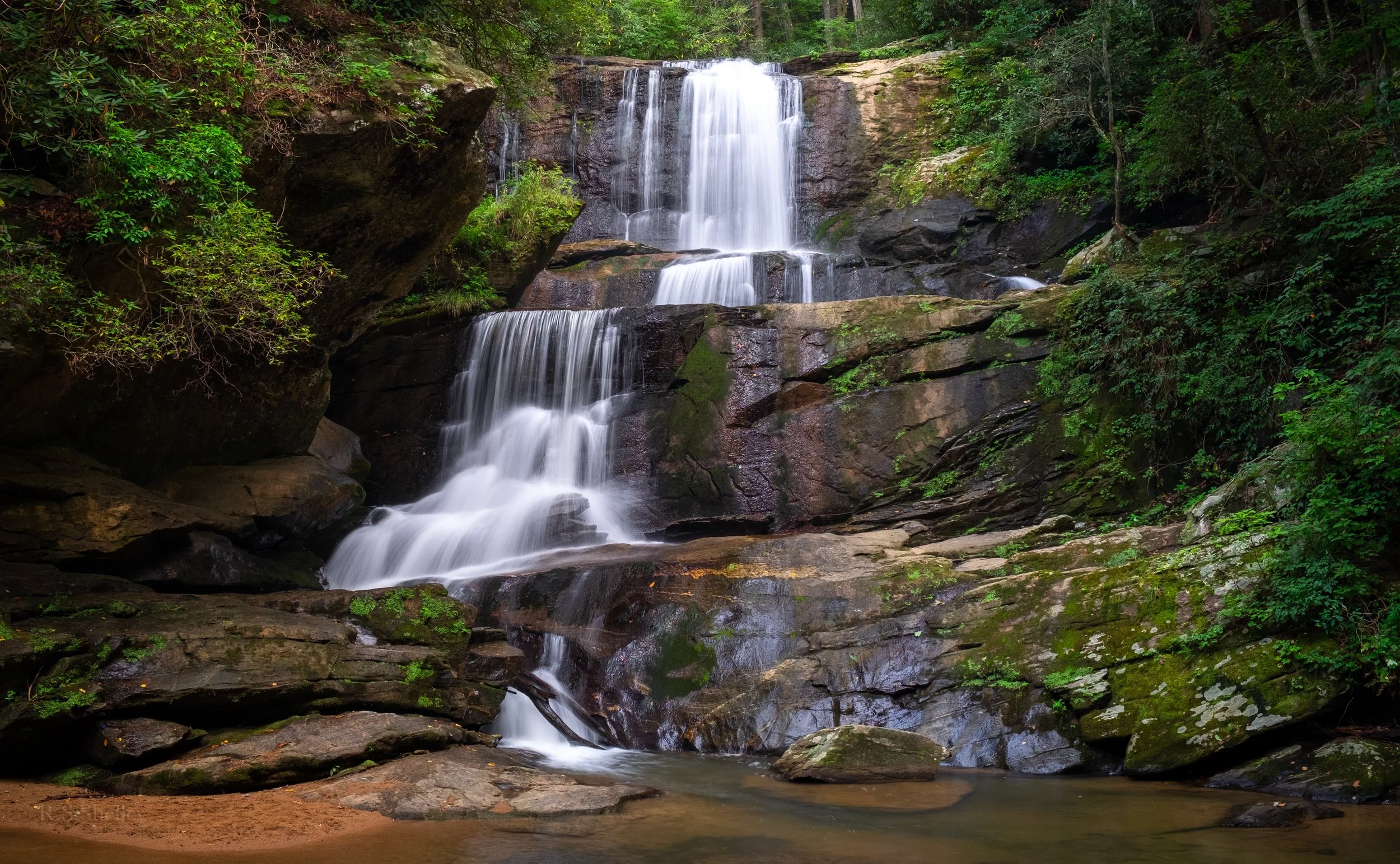 A multi-tiered waterfall flowing over rocks in a lush green forest.
