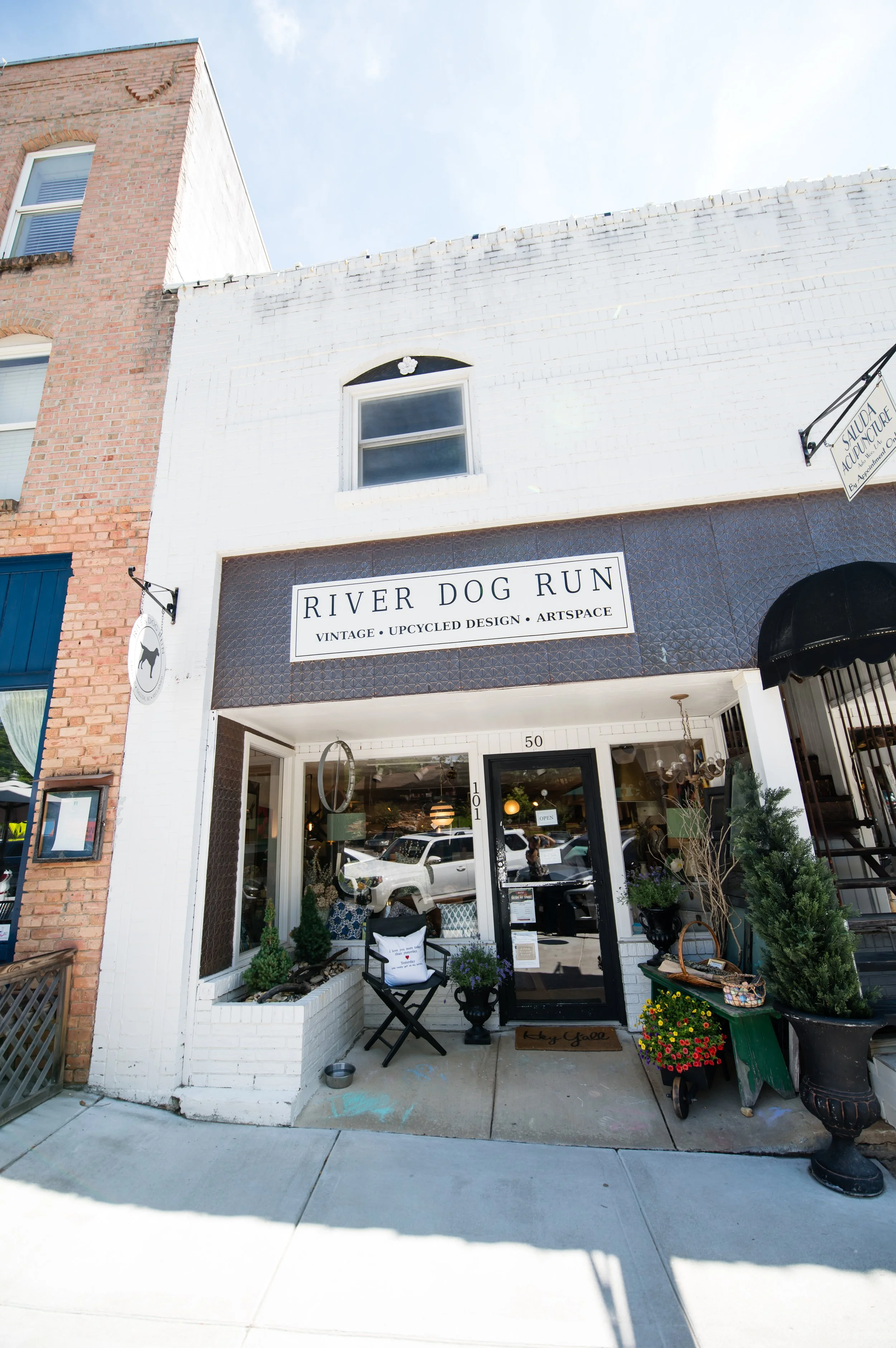  storefront with a sign reading "River Dog Run" and decor including potted plants and outdoor furniture.