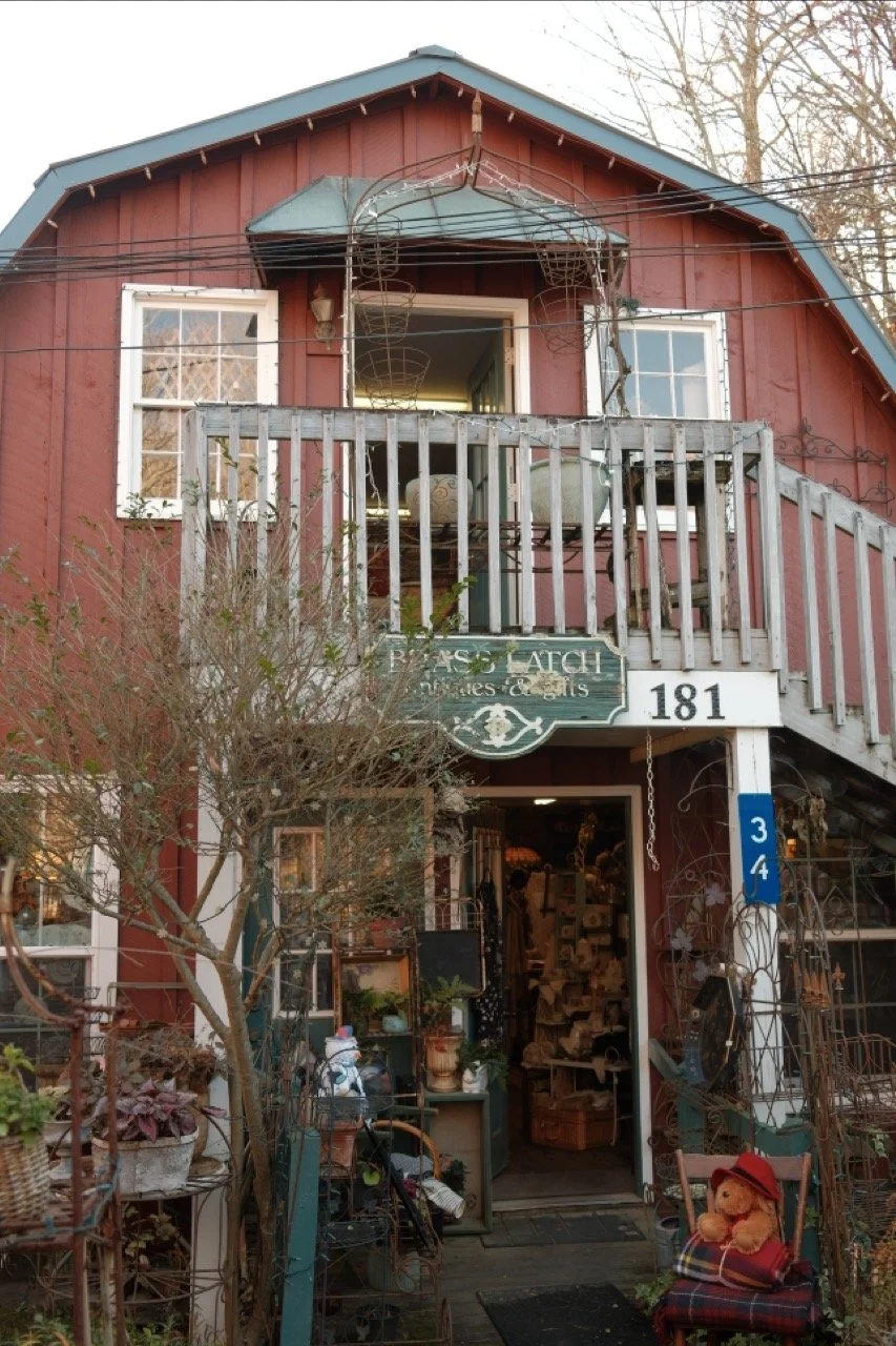 A two-story building with a red wooden exterior and a blue roof, featuring a small balcony on the second floor with gardening supplies and chairs. The first floor is an antique shop with various decorative items, plants, and a teddy bear sitting on a