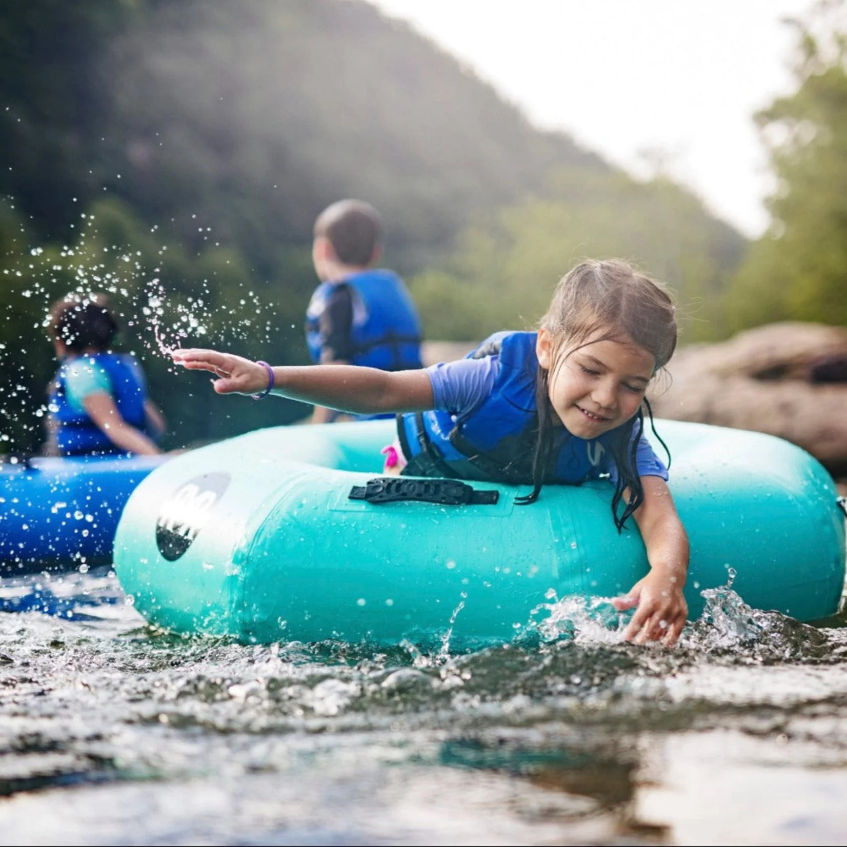 Children enjoying a day by the water, with one girl sitting on a turquoise inflatable ring in the water, splashing and smiling, while others stand in the background.