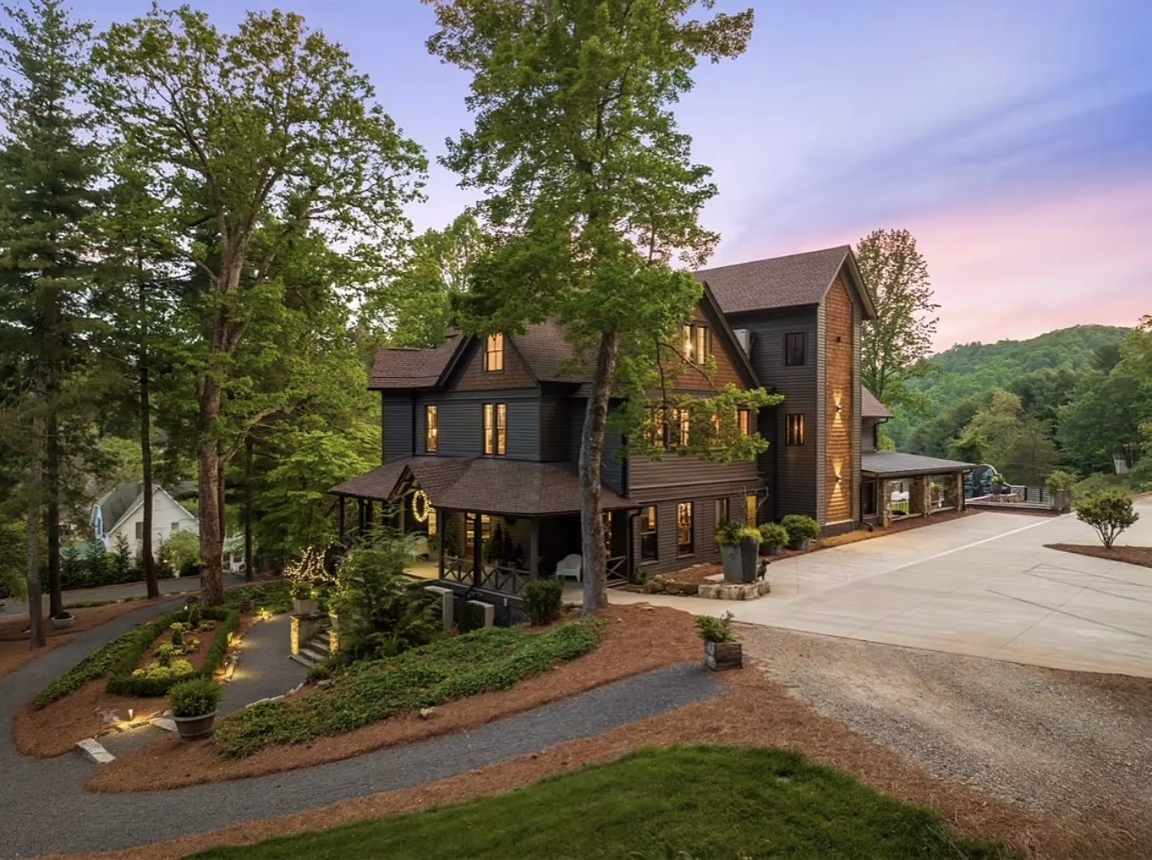 A large, dark-colored house in a wooded area during dusk, with warm lights glowing from inside, surrounded by trees, a front porch, and landscaped yard with potted plants and pathway lights.