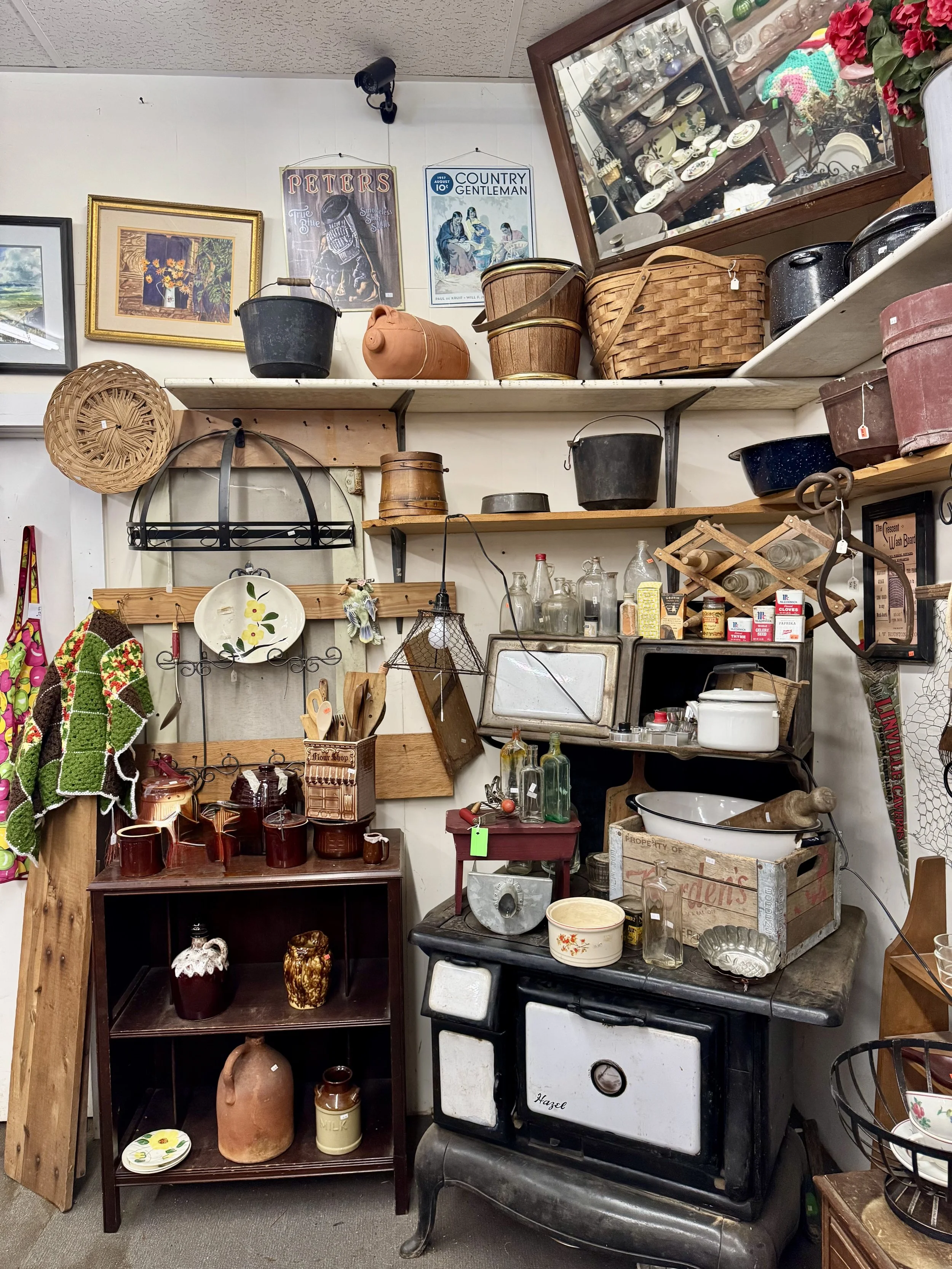 A thrift store display with shelves holding vintage and antique kitchenware, including pots, pans, bowls, and glass bottles, alongside framed artwork and decorative items.