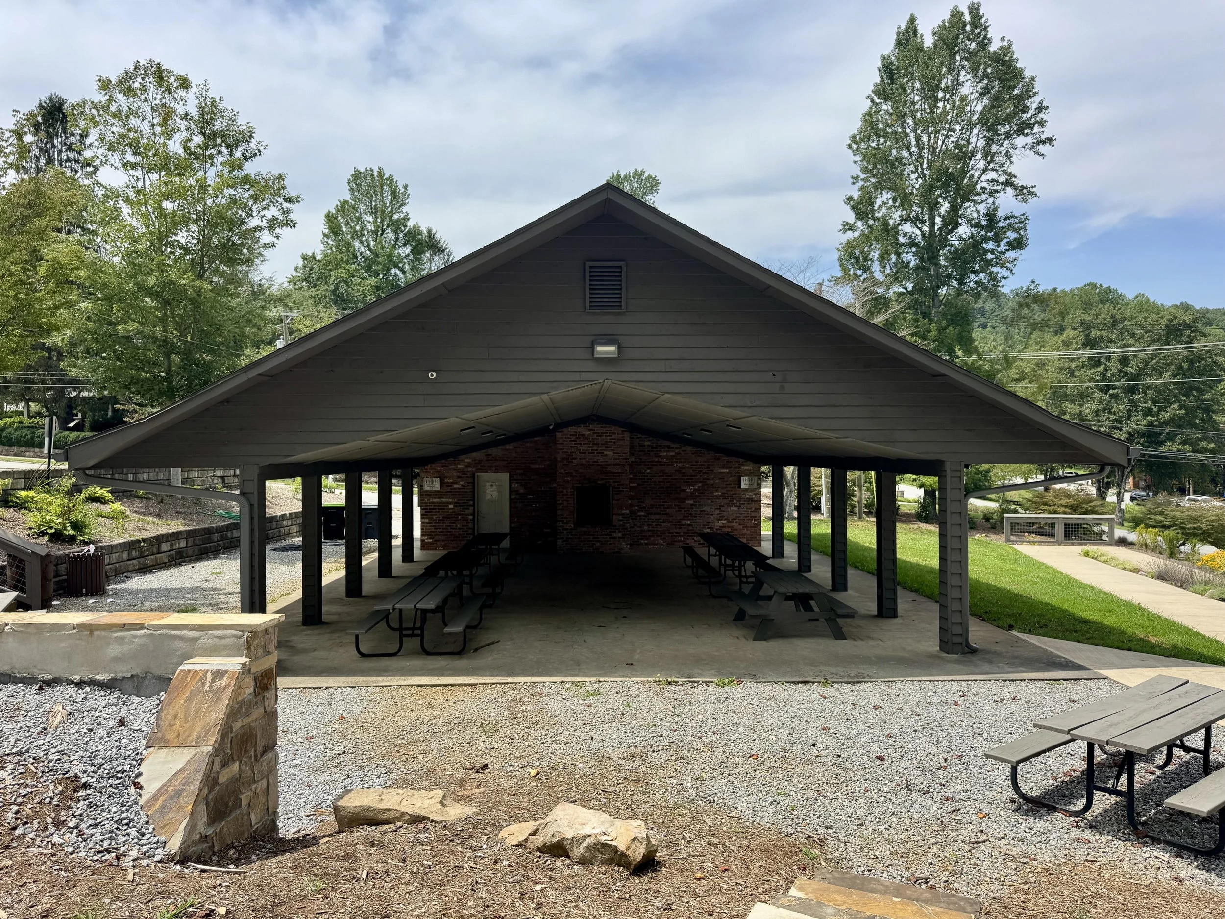 A covered outdoor picnic area with several picnic tables, surrounded by trees and greenery, under a partly cloudy sky.