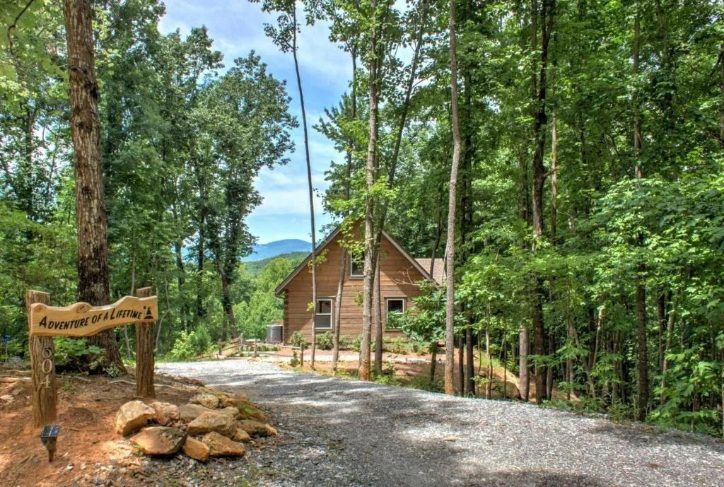 A wooden cabin surrounded by trees and a gravel driveway with a wooden sign that says "Adventure of a Lifetime."