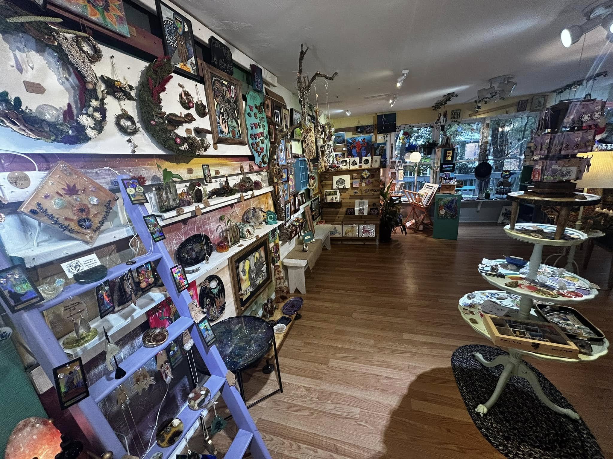 Interior of an art and craft shop with shelves and tables displaying jewelry, paintings, sculptures, and various decorative items. Glass display cases, framed art, and natural wood accents are visible, with a large window letting in natural light.