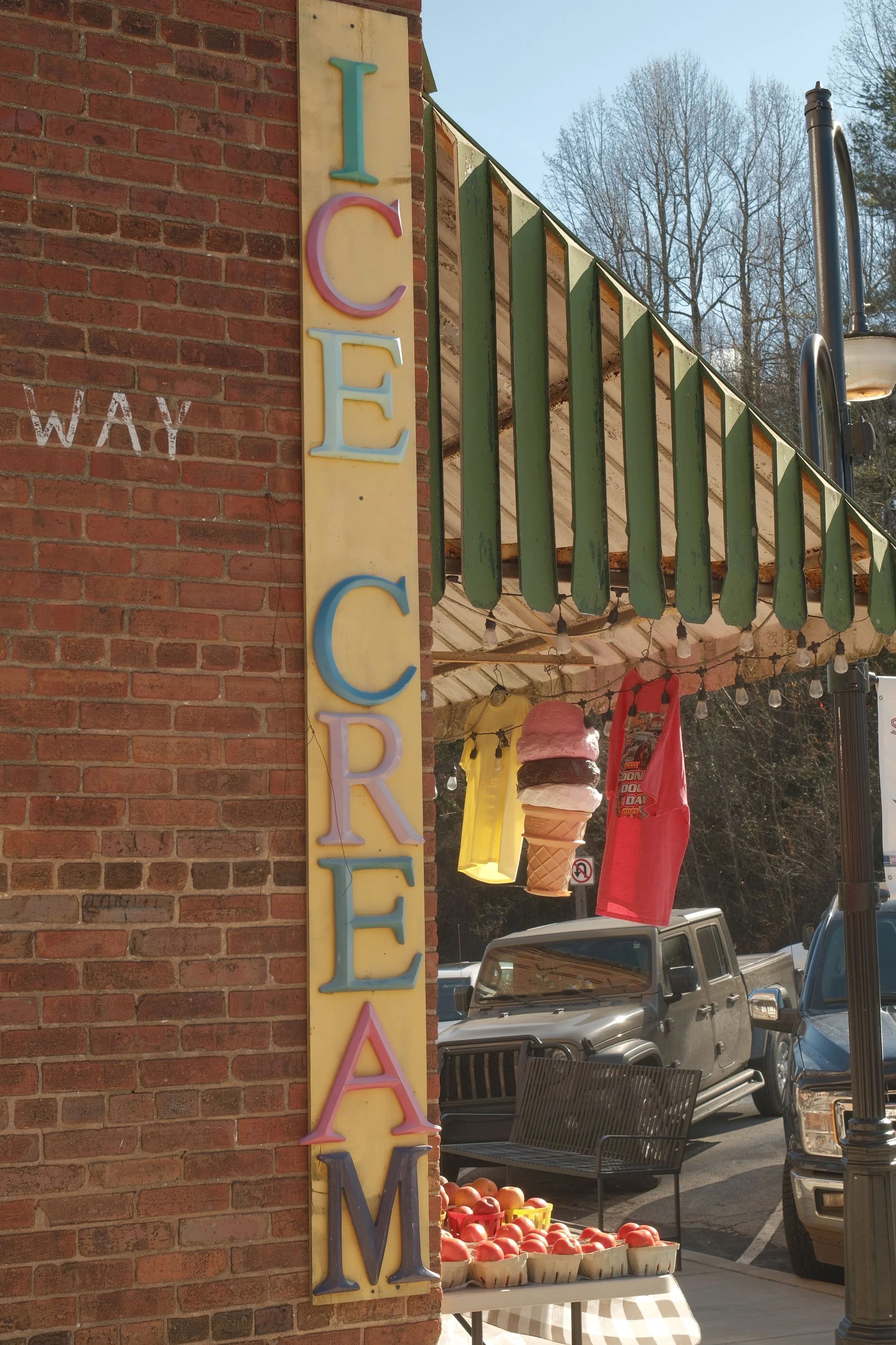 Colorful ice cream sign on brick wall with striped awning, tomatoes displayed on a table, parked cars, outdoor seating, and leafless trees in the background.