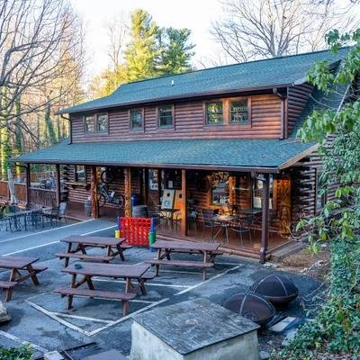 A two-story wooden cabin with a large porch, located in a wooded area, with patio furniture and a ping pong table outside.
