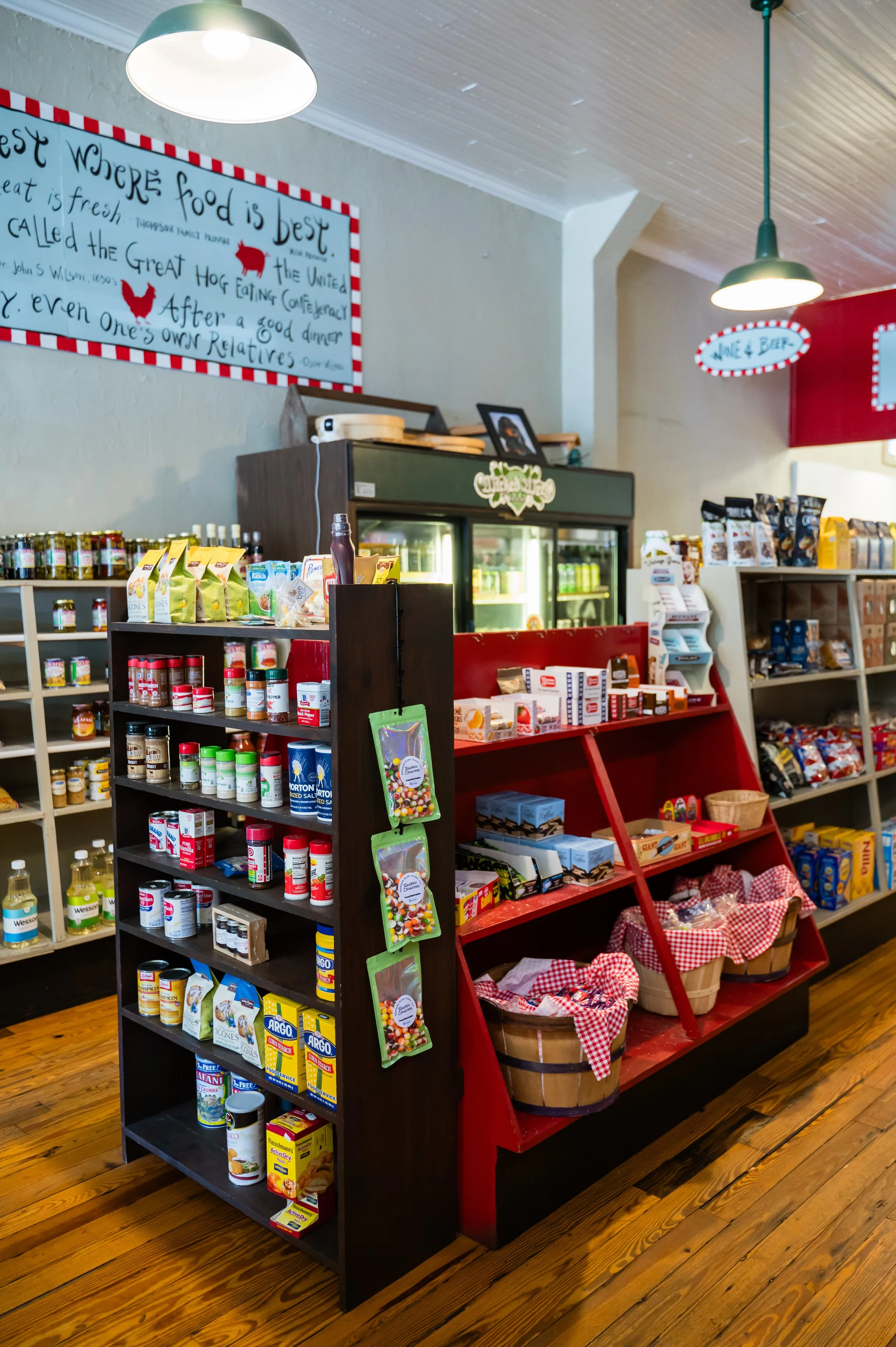 Interior view of a grocery or convenience store with shelves of food items, a display of canned goods, and a red shelf with baskets of baked goods. There are pendant lights hanging from the ceiling and a large decorative sign on the wall with a quote