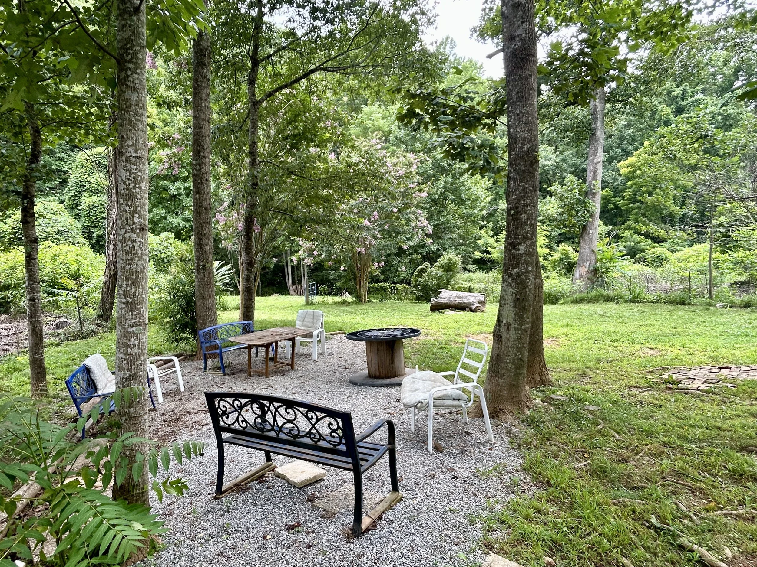 A backyard scene with multiple trees, green foliage, and outdoor furniture including chairs and a table on a gravel ground.