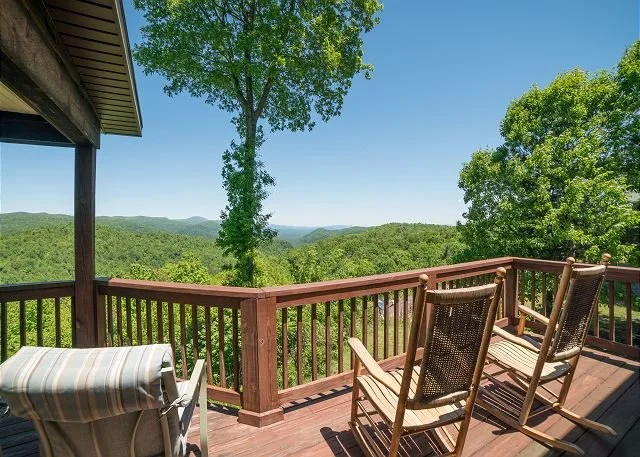 View of a wooden deck with outdoor chairs overlooking a green forested landscape and distant mountains under a clear blue sky.