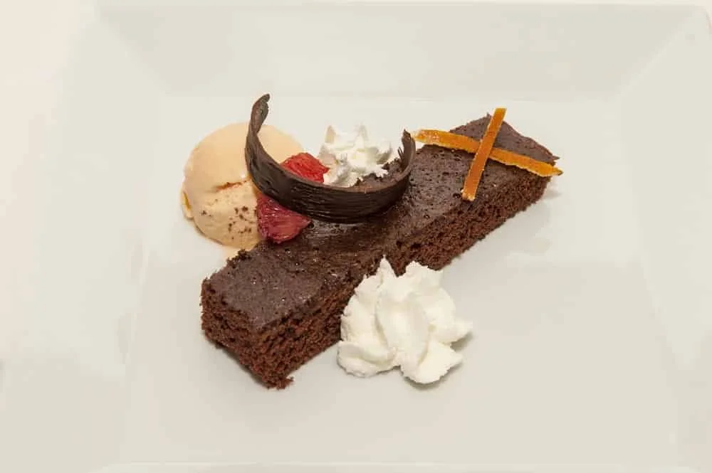 Chocolate cake slice with whipped cream, ice cream, chocolate curl, dried strawberry, and orange zest garnishes on a white plate.